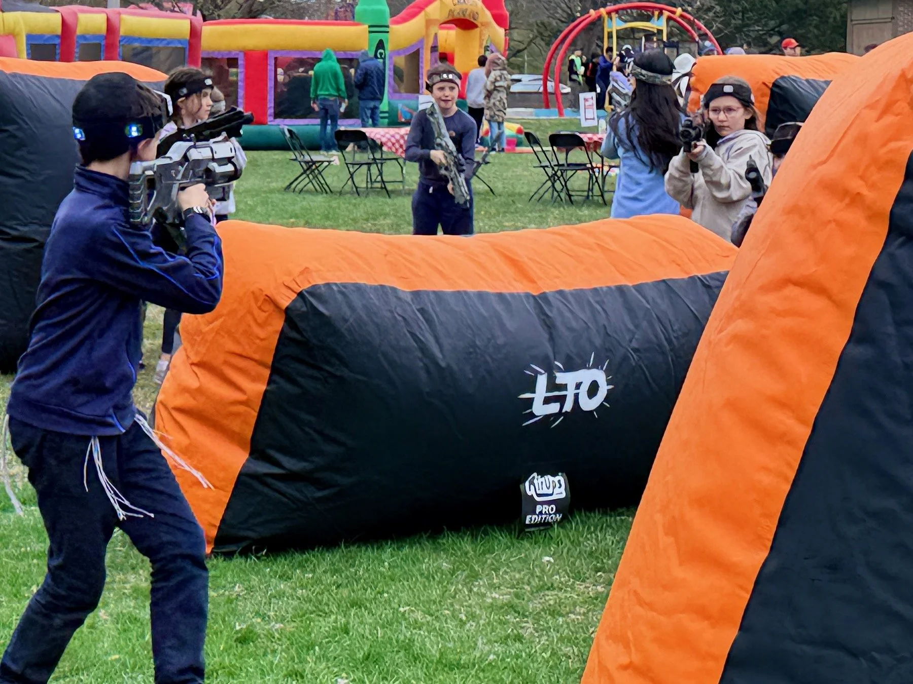 Children playing paintball outdoors on a grassy field with inflatable obstacles, wearing protective gear and holding paintball guns, with a colorful inflatable playground in the background.