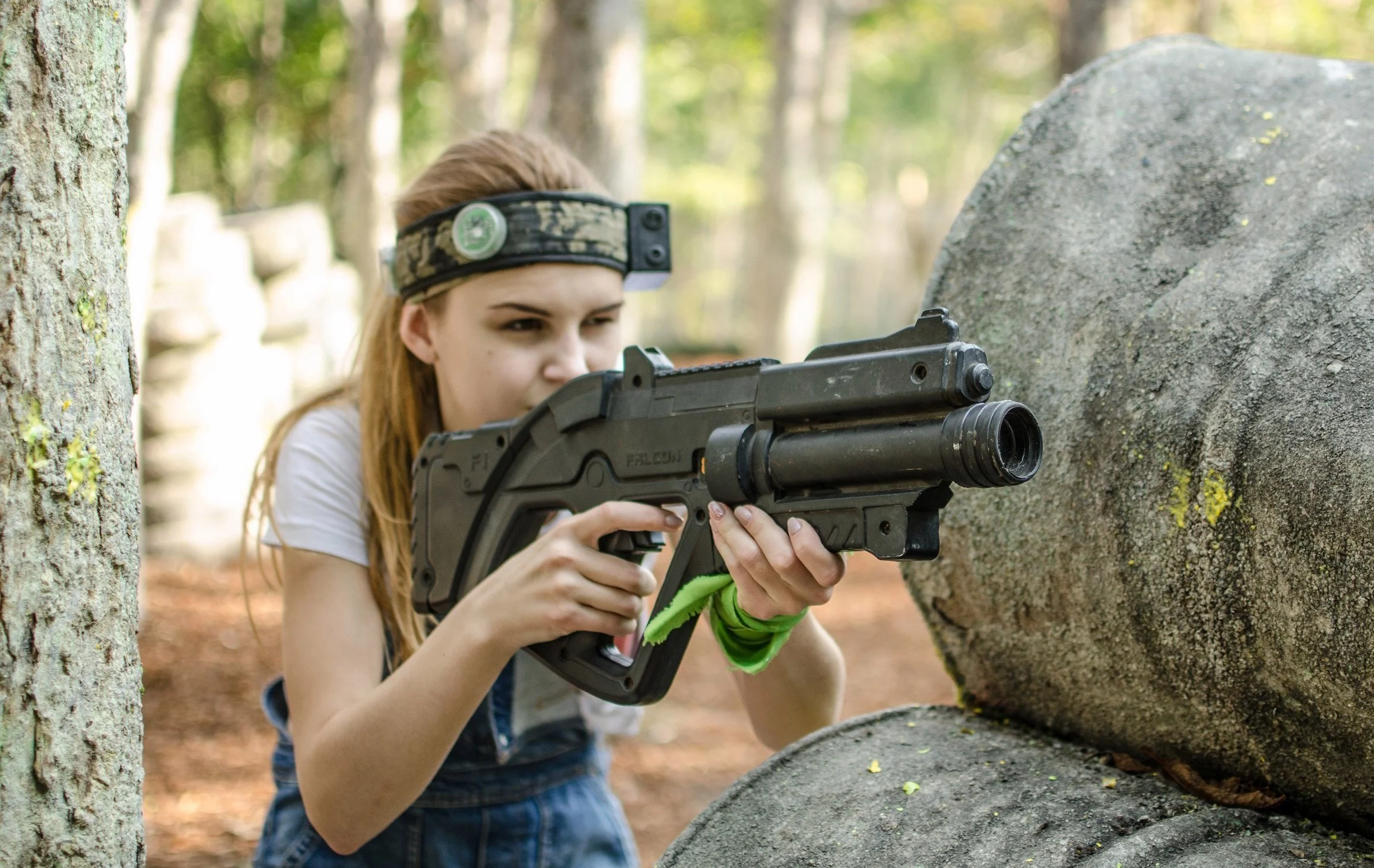 Young girl in a headlamp aiming a toy gun in a wooded area.