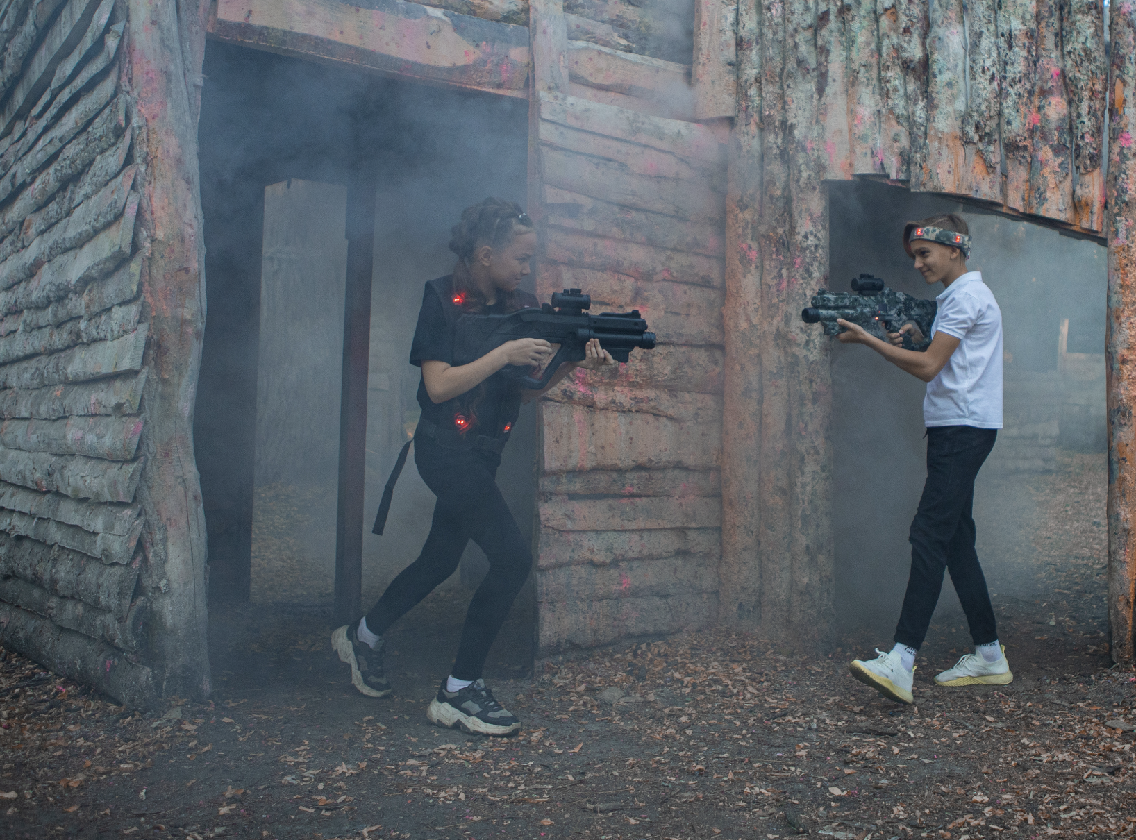 Two young girls playing laser tag outdoors, holding laser guns, with a smoky atmosphere and a dilapidated building in the background.