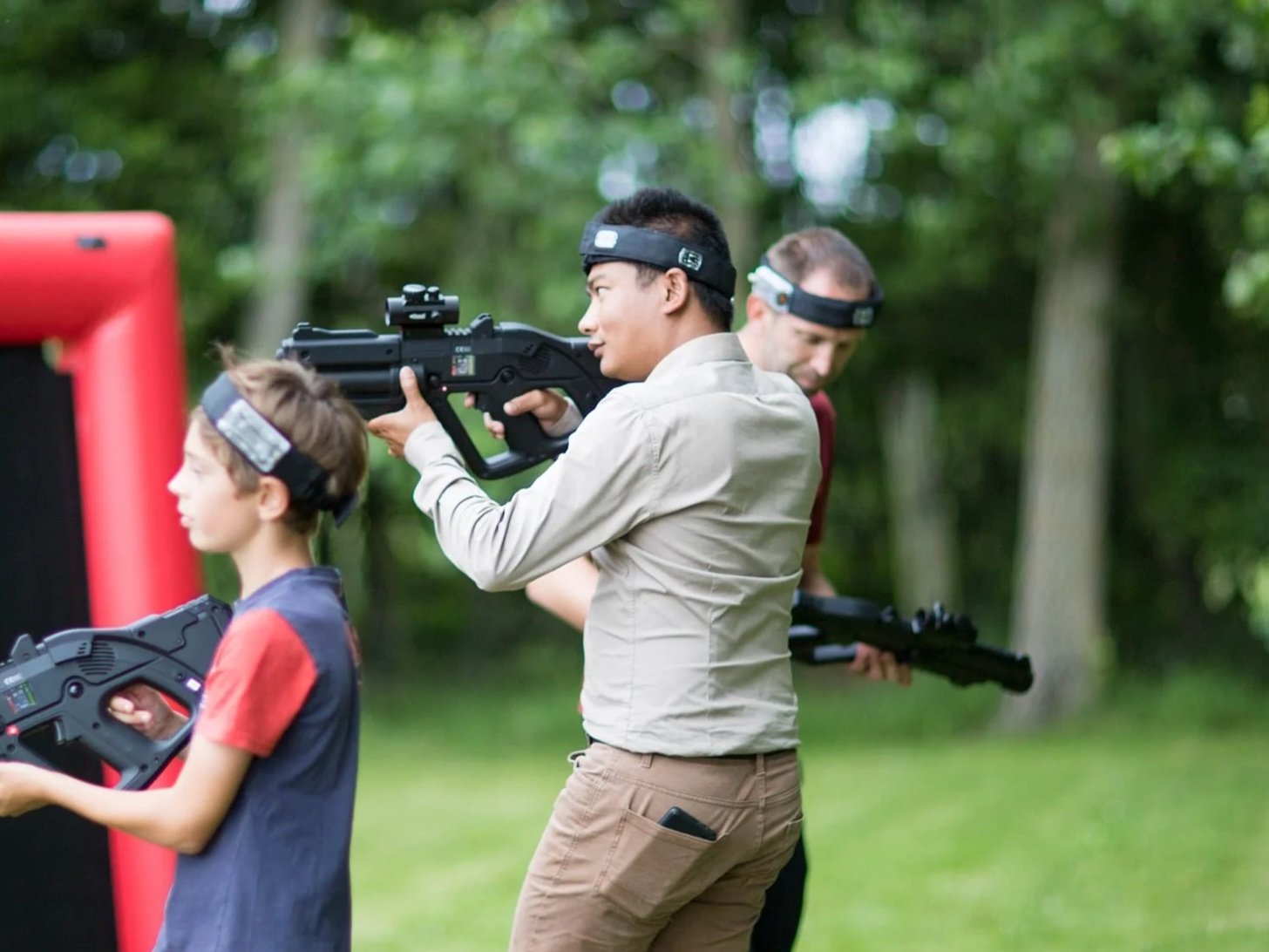 A group of people engaged in laser tag outdoors, with two men and a boy all holding laser tag guns and wearing headbands.
