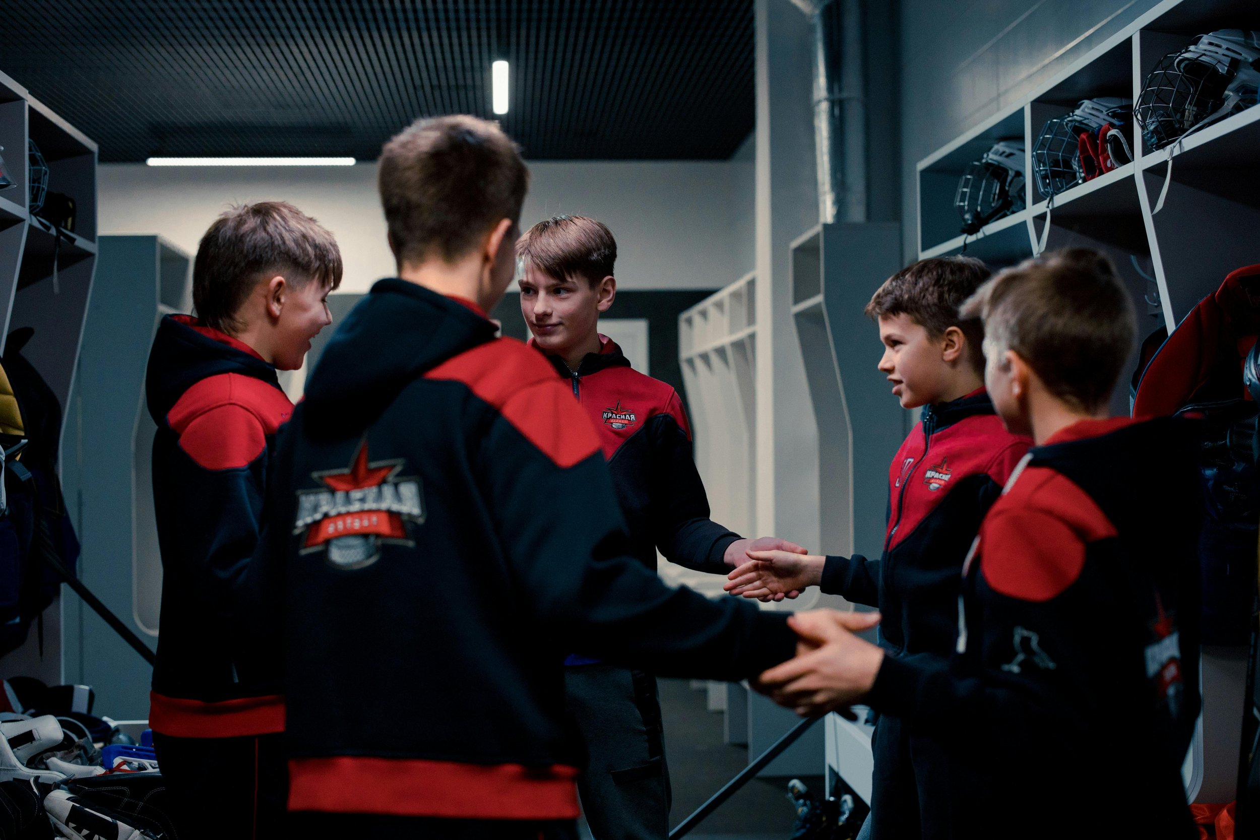 A group of young boys in red and black hockey uniforms in a locker room, shaking hands and talking.