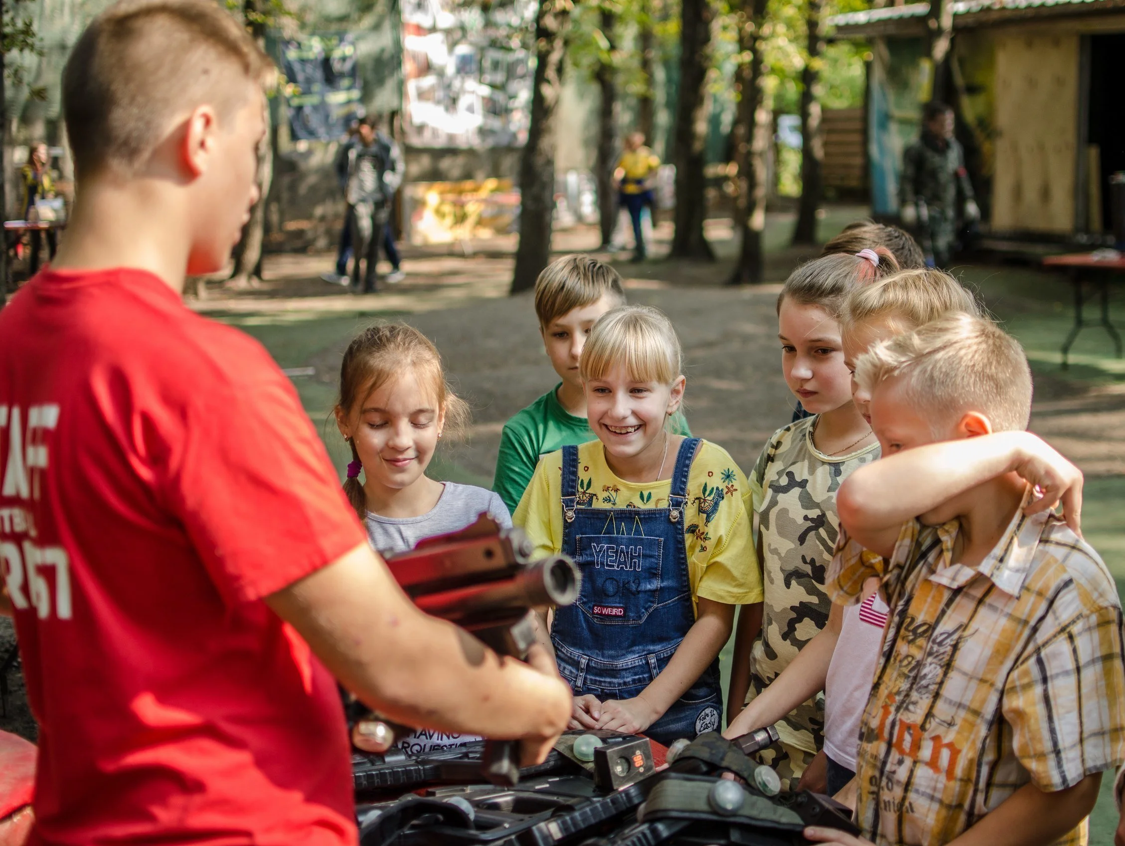 A group of children gathered around a person holding a toy gun, outdoors in a wooded area, with trees and other people in the background.