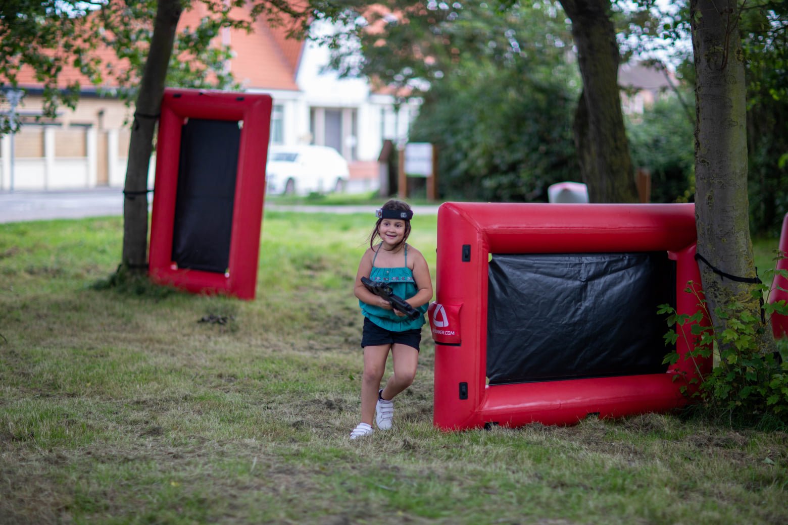 A young girl smiling while playing laser tag outdoors, holding a laser gun, with inflatable barriers around her and trees in the background.