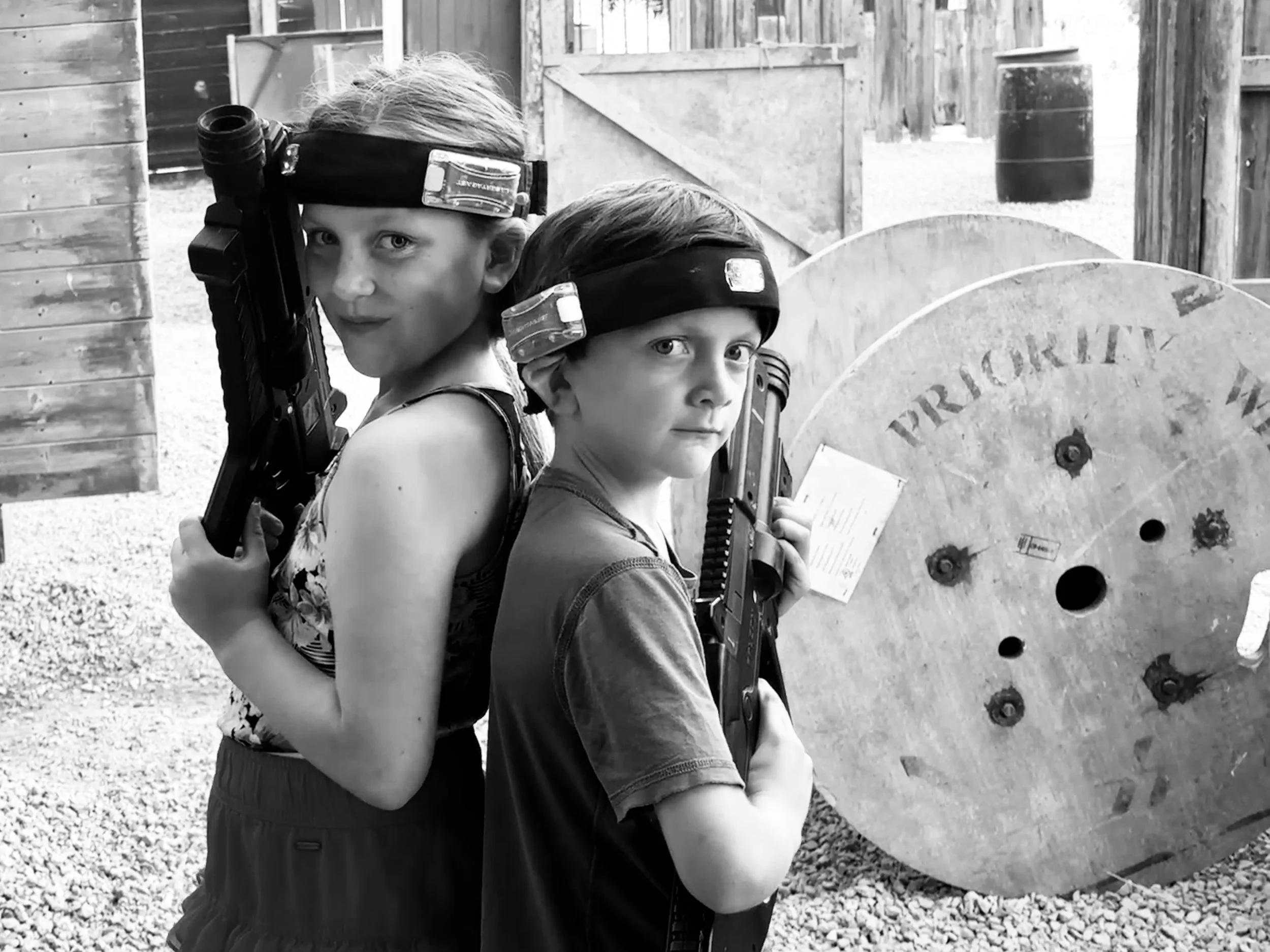 Two children with toy guns and headbands posing in front of a wooden fencing at a paintball or laser tag game area.
