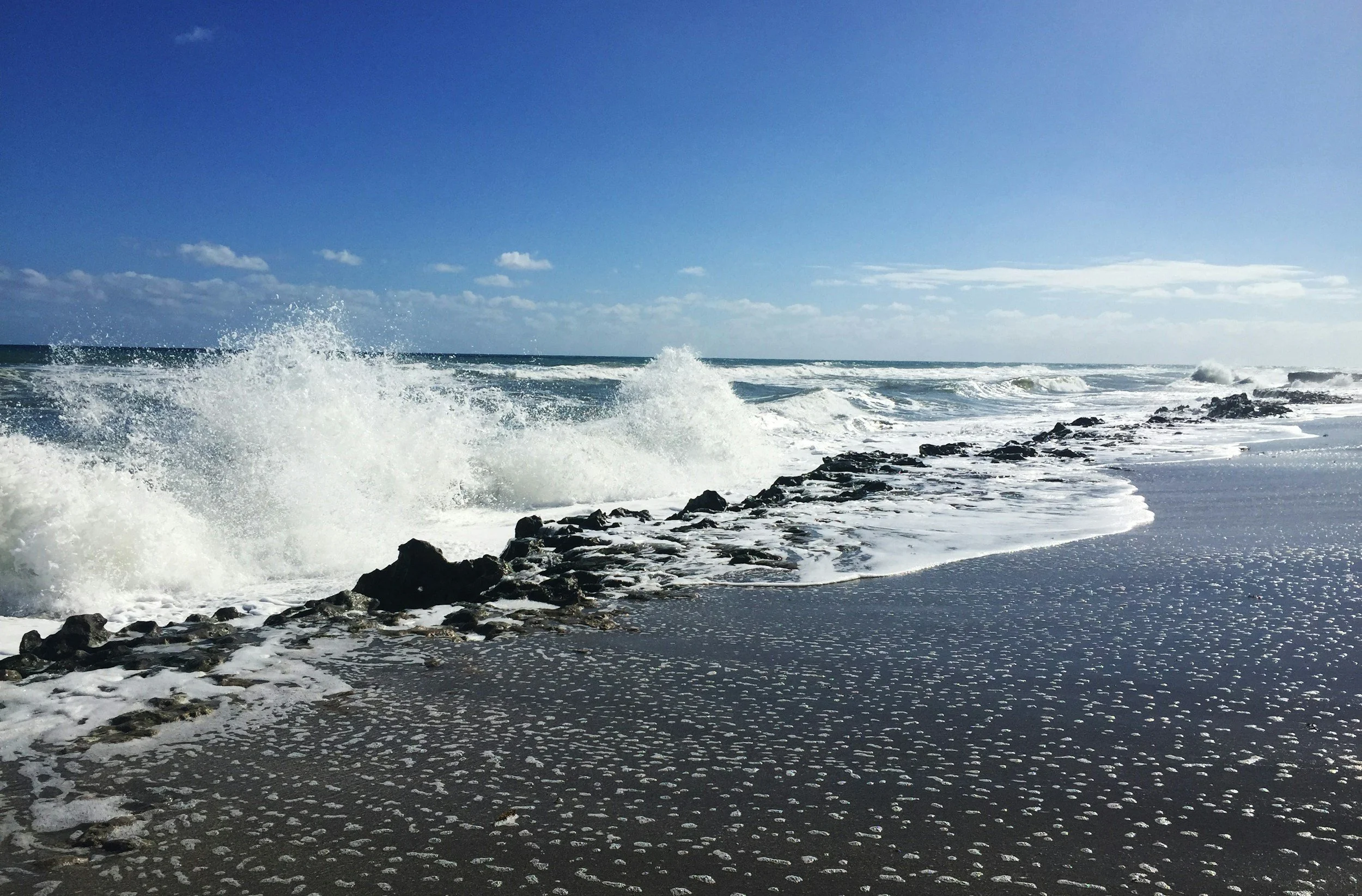 Ocean waves crashing onto a rocky shoreline under a clear blue sky with some clouds.