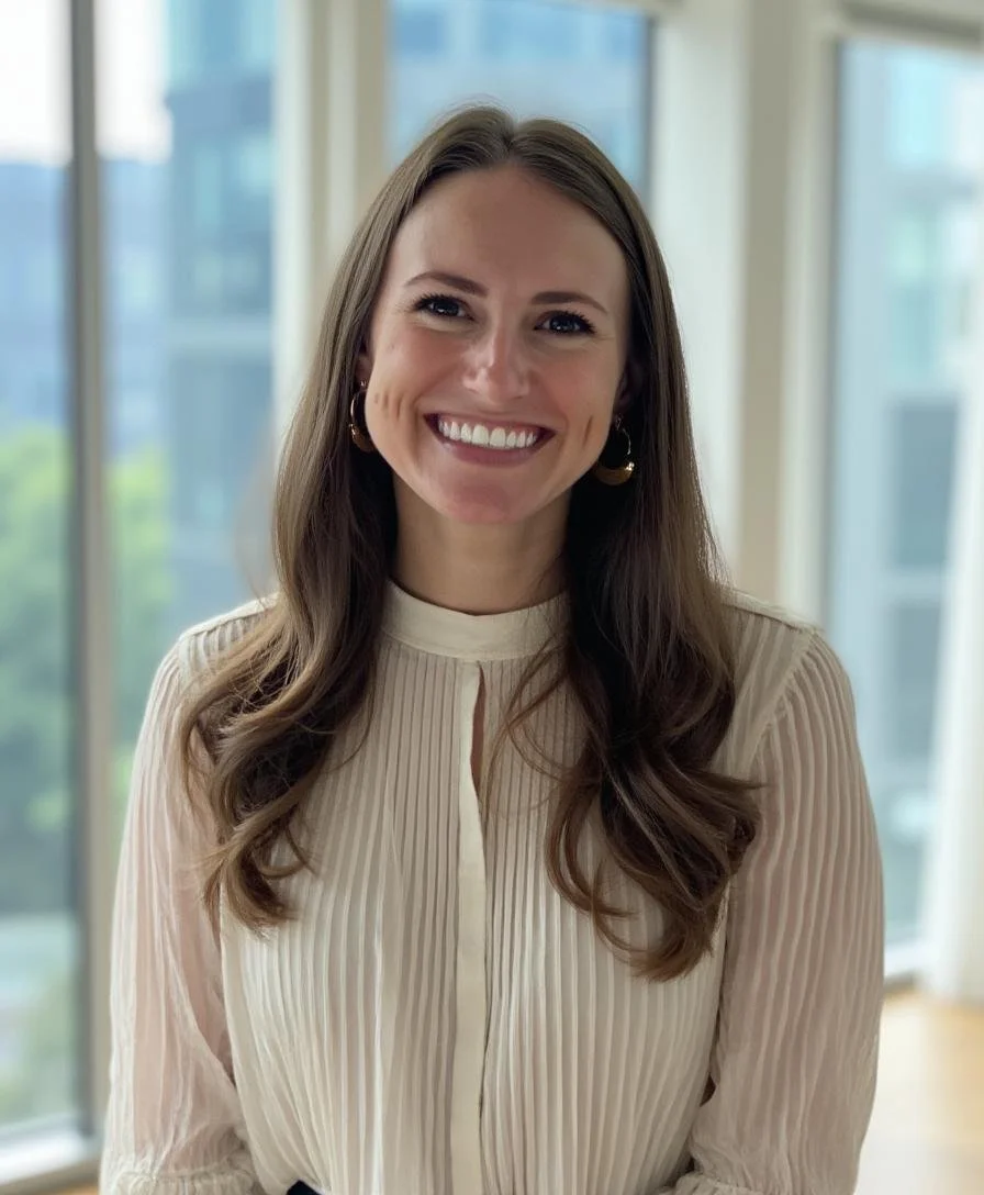 A woman with long brown hair, smiling, wearing a cream-colored pleated blouse and hoop earrings, in a bright indoor setting with large windows.