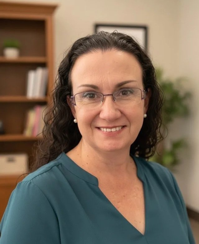 A woman with long dark curly hair, glasses, and pearl earrings smiling at the camera indoors with a bookshelf and framed picture in the background.
