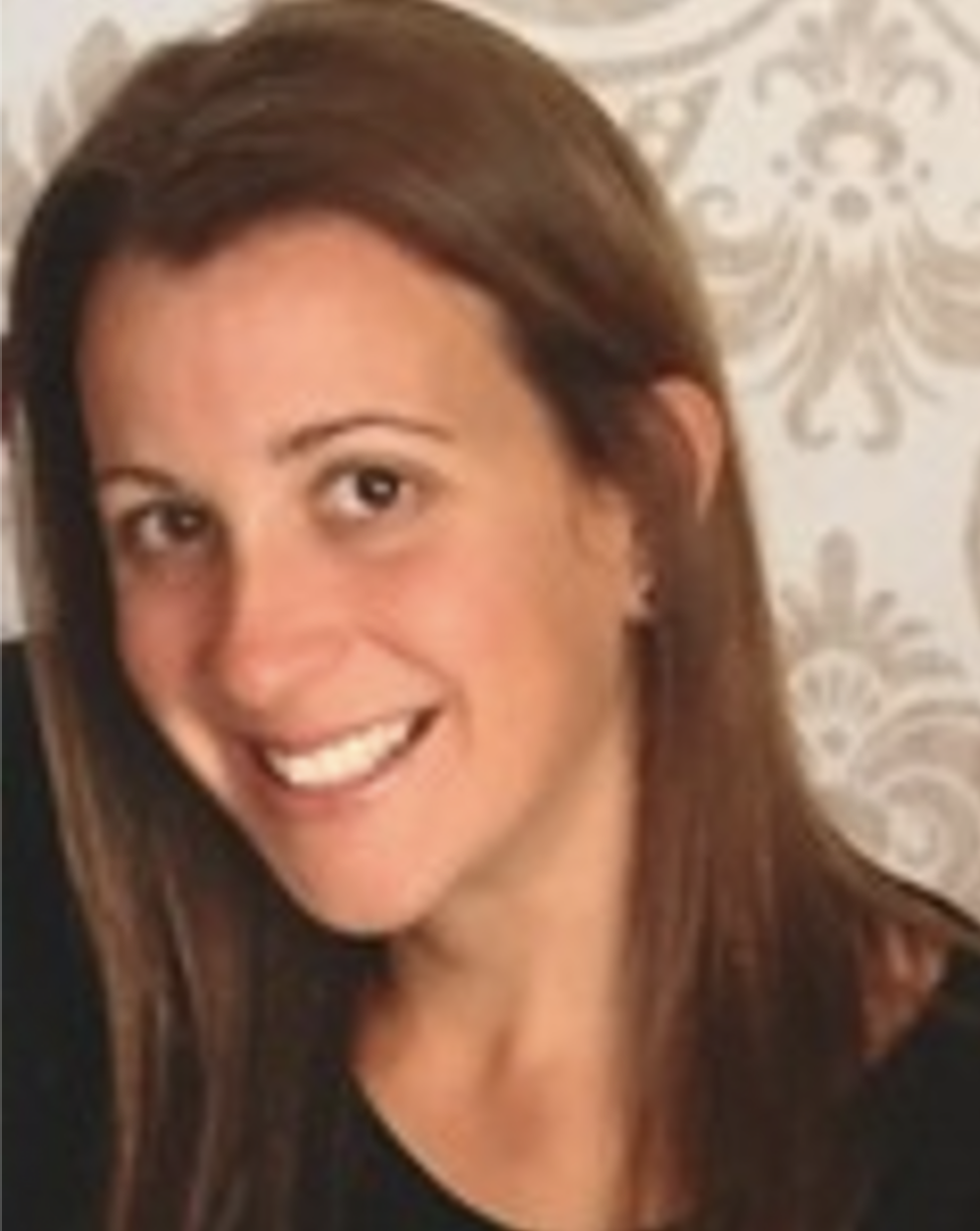 Close-up of a smiling woman with brown hair and light skin, wearing a black top, in front of a patterned wallpaper background.