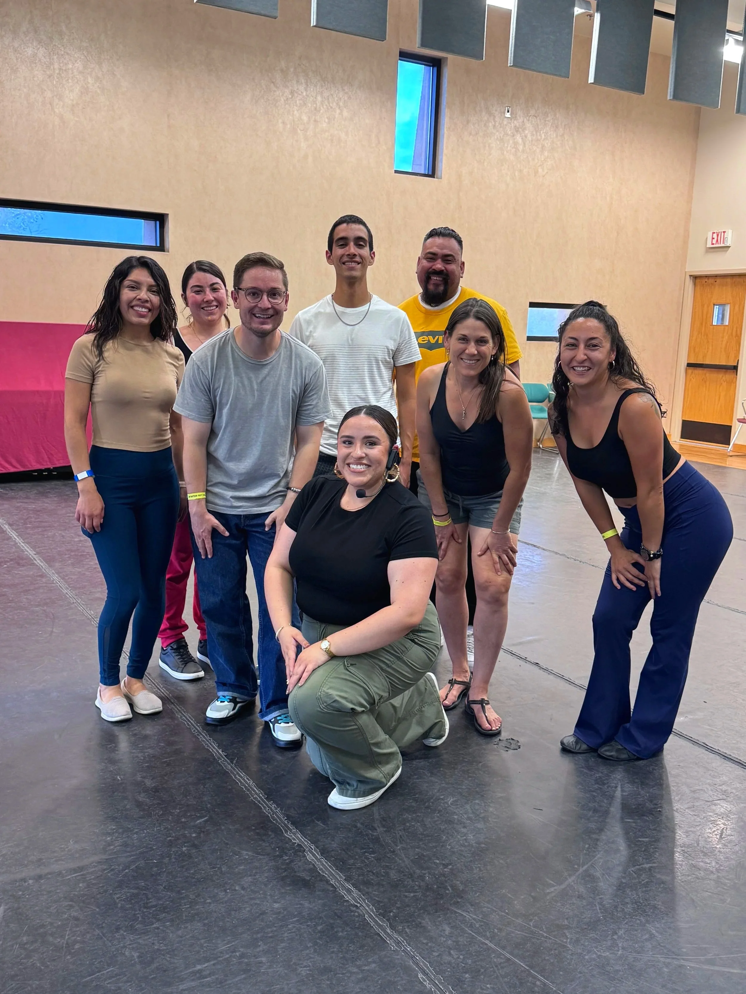 Group of eight people smiling and posing together in a spacious room with beige walls and high ceilings.