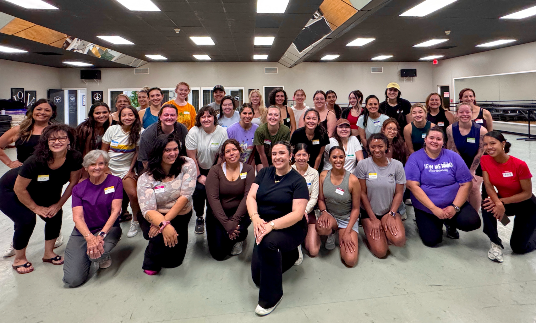 Group of diverse women smiling in a dance studio with mirrors on the ceiling and walls, some kneeling and some standing, in a casual setting.