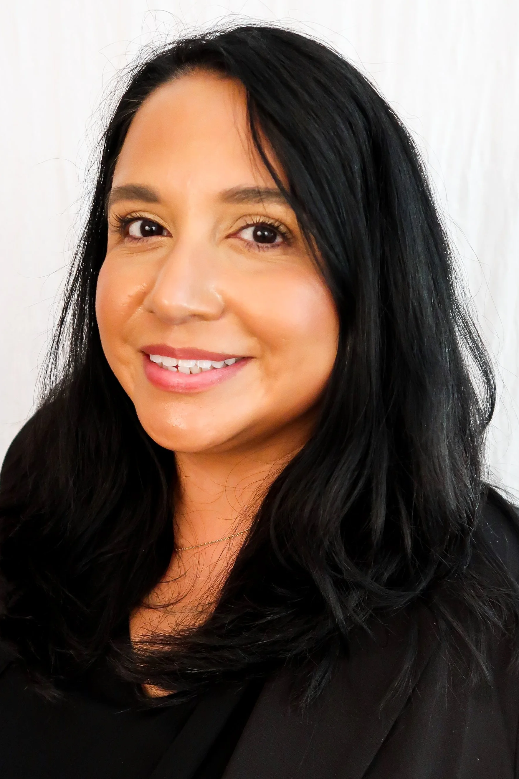 A woman with dark hair smiling and wearing a black blazer and top, standing against a white background.