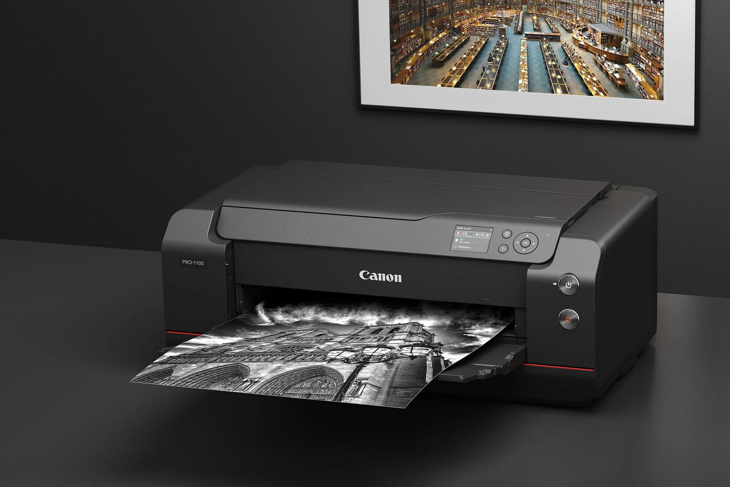 A black Canon imagePROGRAF printer printing a black and white architectural photograph on a dark desk with a framed art print of a library on the wall behind it.