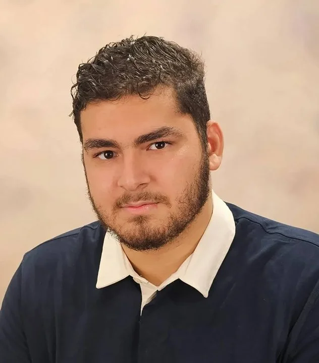 A young man with dark, curly hair and facial hair, wearing a dark blue shirt over a white collared shirt, looking at the camera with a neutral expression.