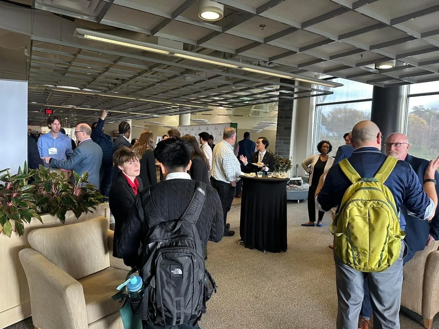 Business conference attendees networking and chatting in a modern indoor venue with large windows, plants, and a high ceiling.