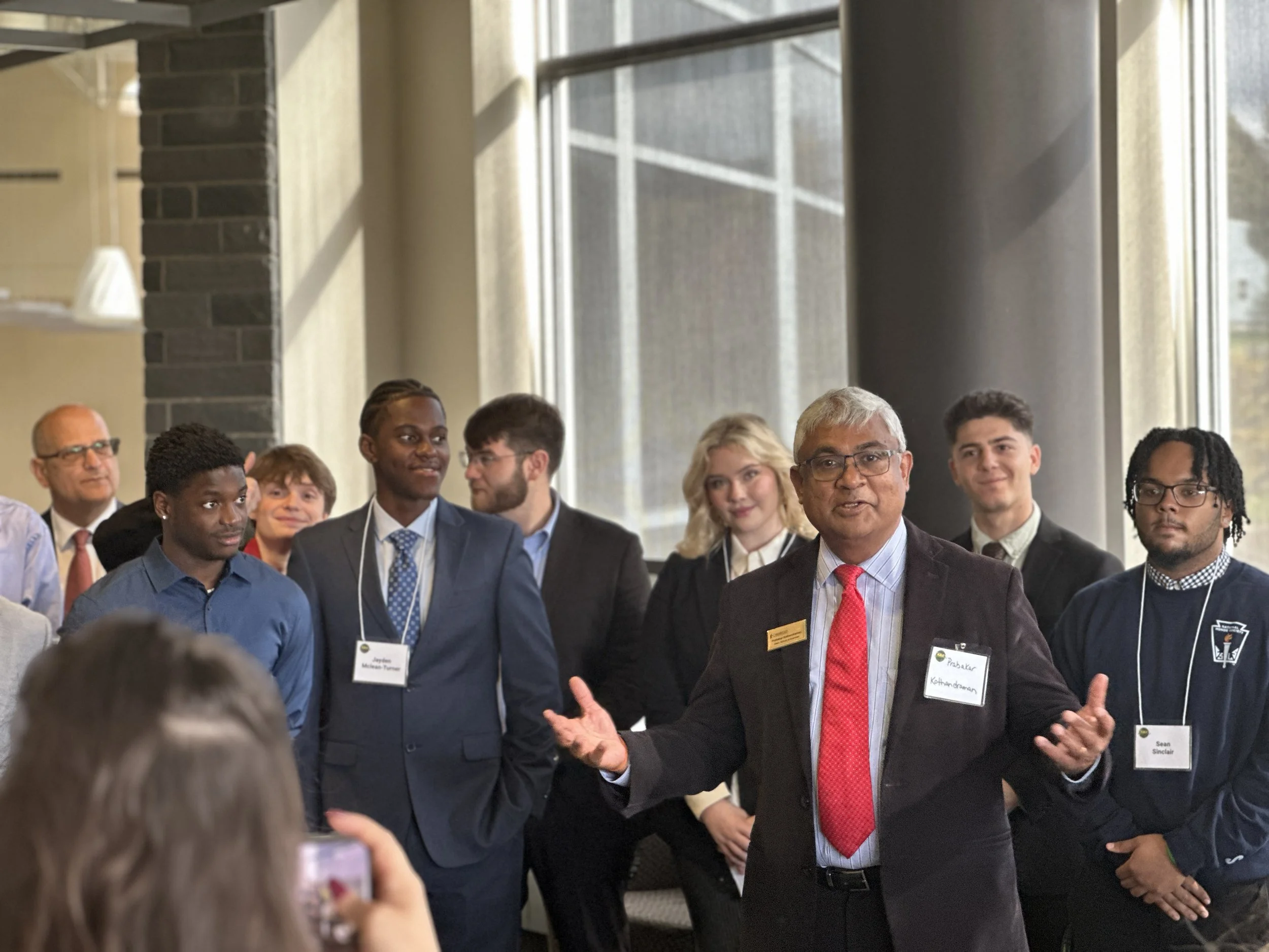 A man with gray hair and glasses speaking to a group of diverse young professionals, some wearing suits and name tags, inside a modern building with large windows.