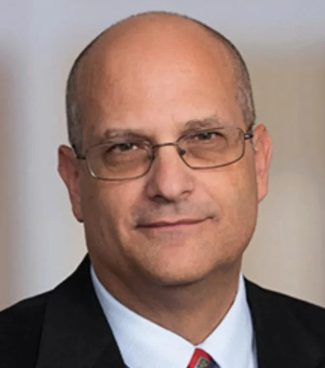 A middle-aged man with glasses, wearing a black suit, white shirt, and a patterned tie, posing for a professional portrait.
