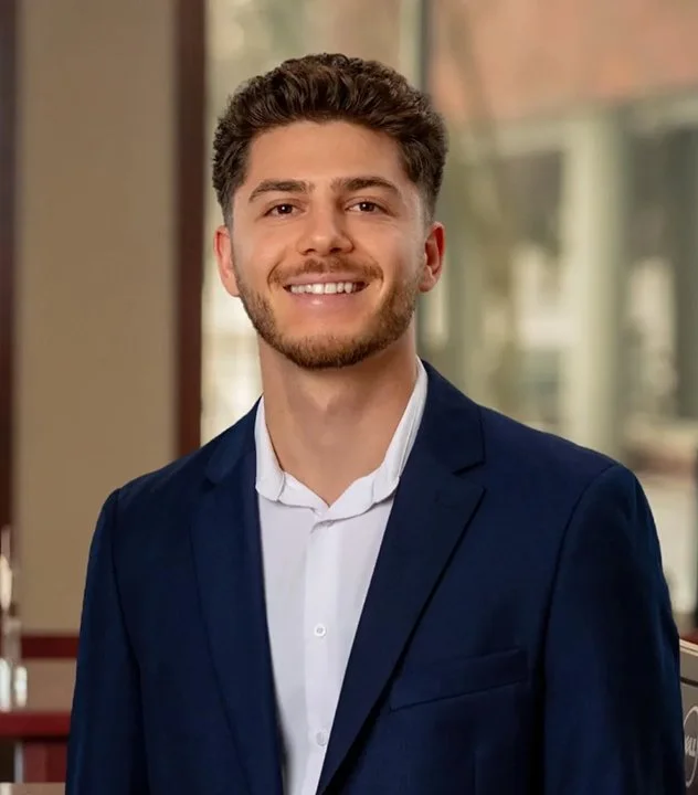 A young man with dark curly hair and a beard, smiling, wearing a navy blue suit and white shirt, in a well-lit indoor setting with large windows in the background.