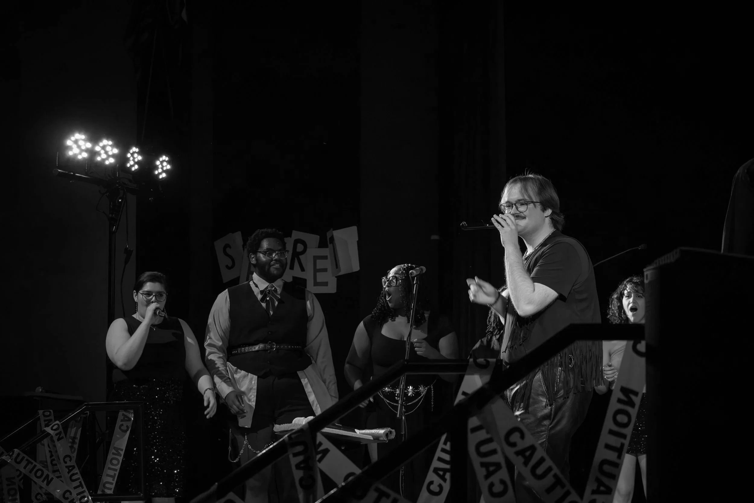 Group of six performers on stage, singing into microphones, with caution tape in the foreground, in a dark setting with stage lighting.