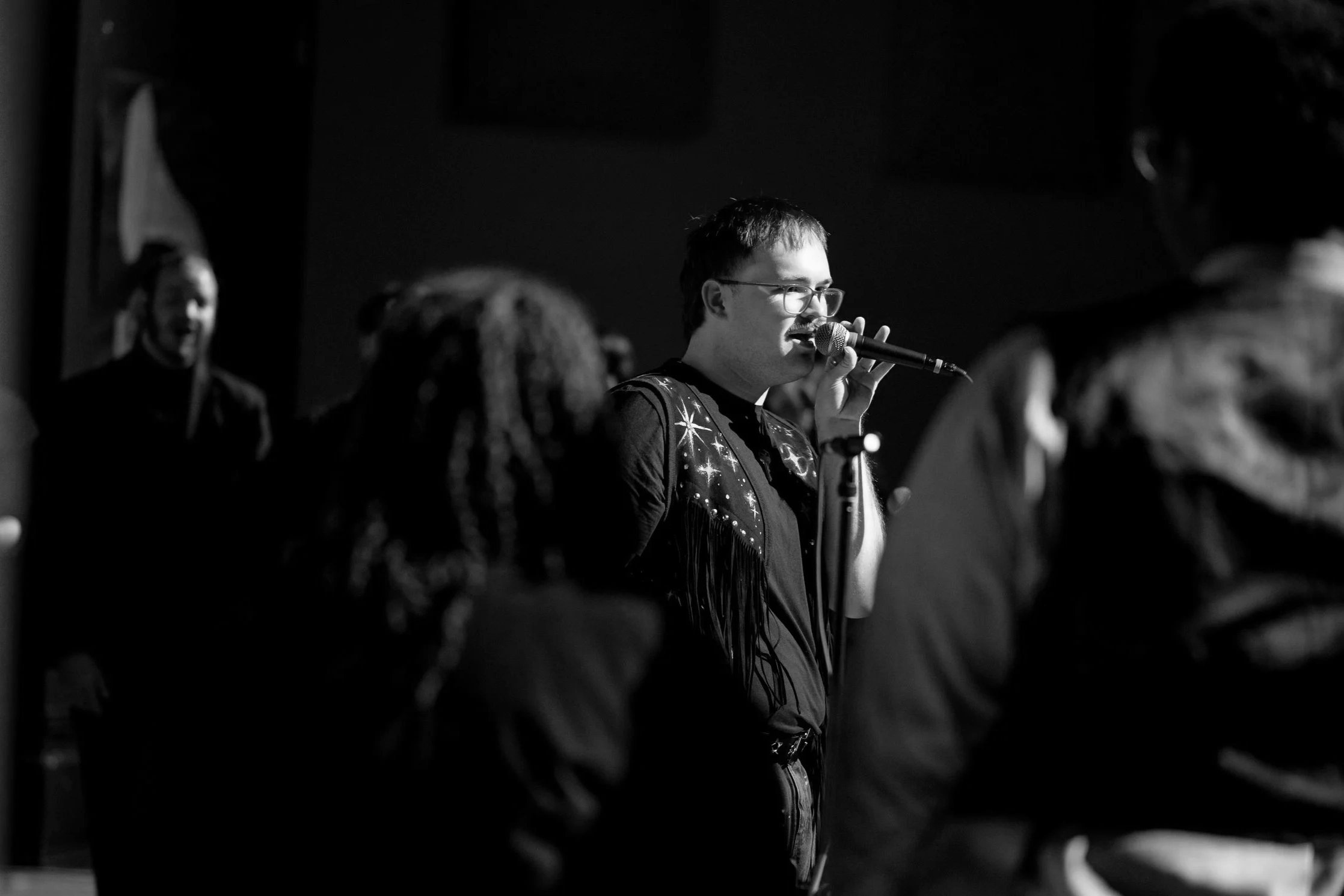 William Reeves wearing a fringe vest singing into a microphone in a dimly lit room, with several people standing nearby.