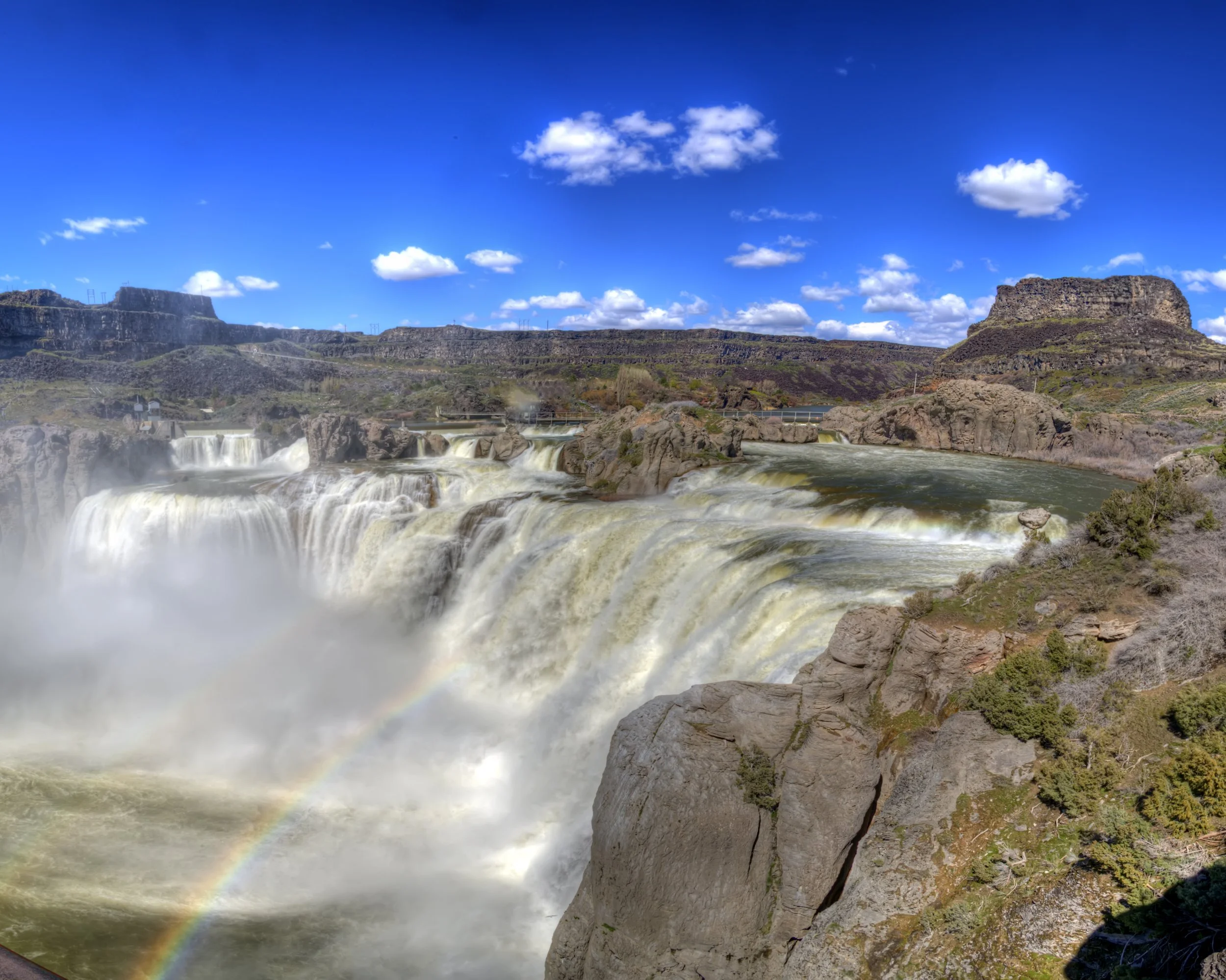 A wide view of a waterfall with multiple cascades, rocks, and surrounding landscape under a blue sky with white clouds. A rainbow is visible near the bottom of the waterfall.