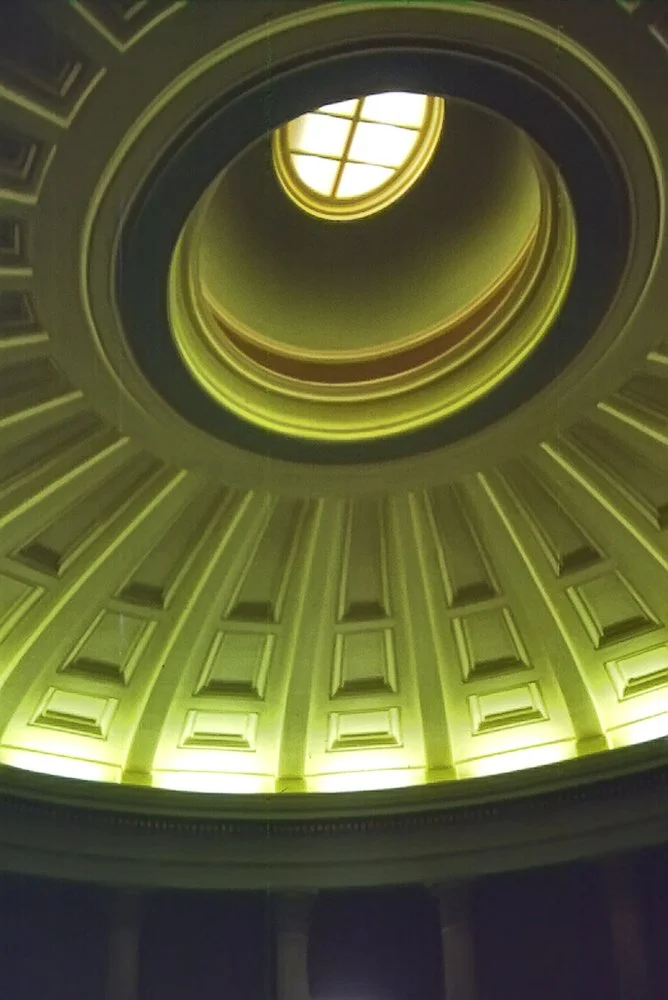 Looking up inside a spiraling staircase with a large round window at the top, illuminated in yellow green light.