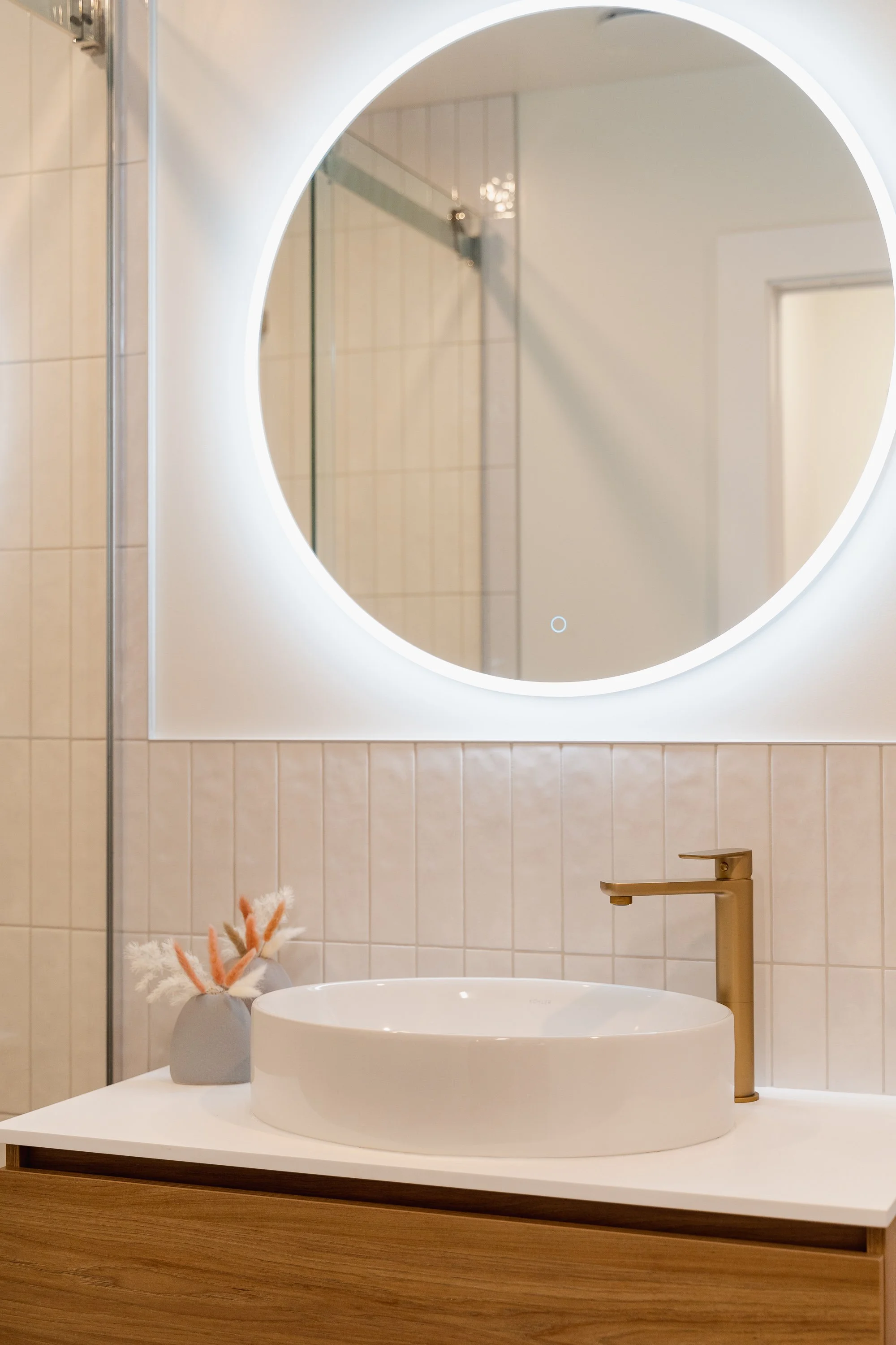 Modern bathroom with a round mirror illuminated by LED backlight, a white vessel sink, and a gold faucet. A small vase with decorative feathers sits on the countertop.