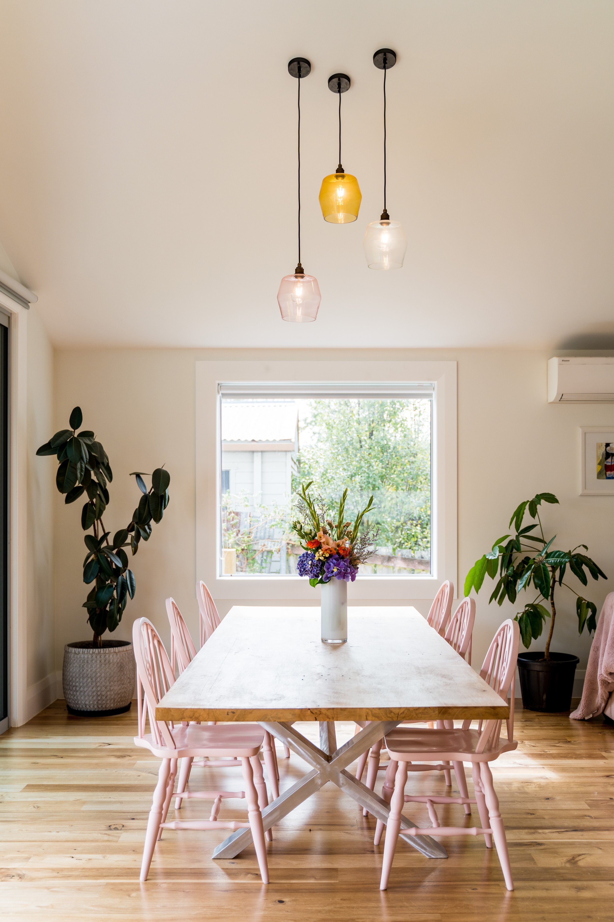 Bright dining room with a window, a wooden table, pink chairs, and floral arrangements, with hanging pendant lights. Passive house principles, high performing, warm and dry. Dunedin, NZ