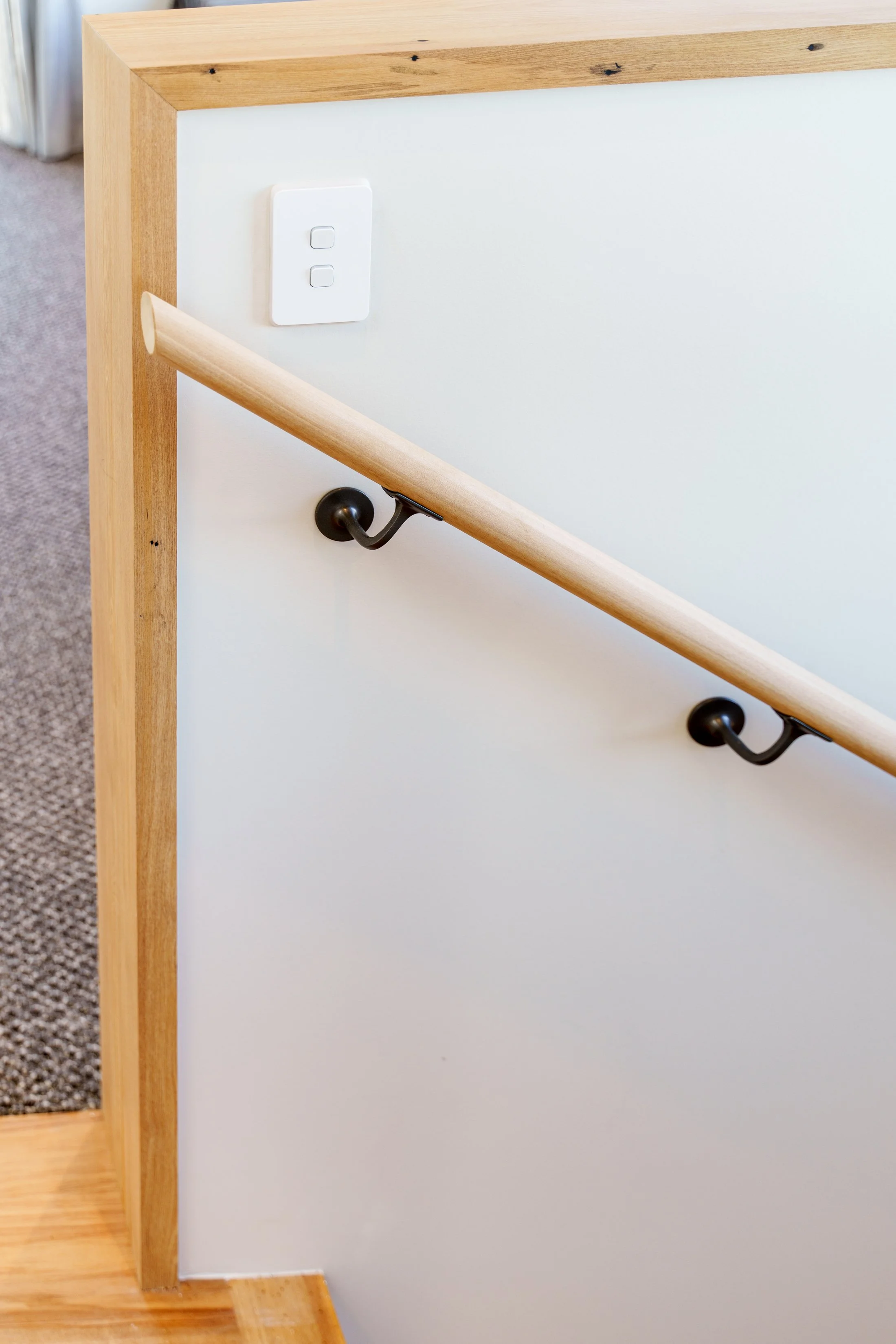Recycled rimu. Passive house principles, high performing, warm and dry. Close-up of a stair handrail made of light wood attached to a white wall with black metal brackets, and a white light switch on the wall to the upper left.