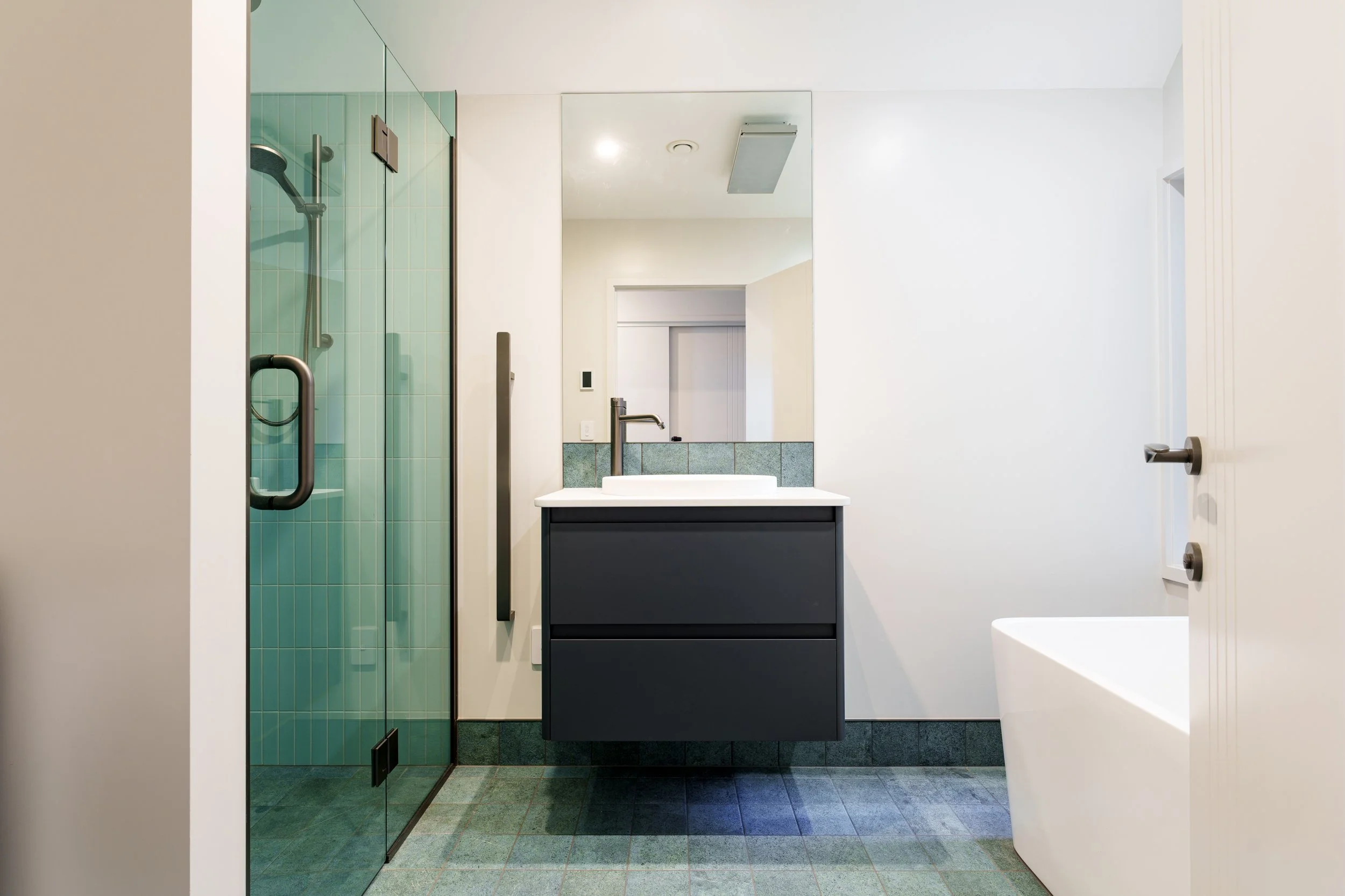 Modern bathroom with teal-tiled shower, dark vanity with vessel sink, large mirror, and freestanding bathtub.