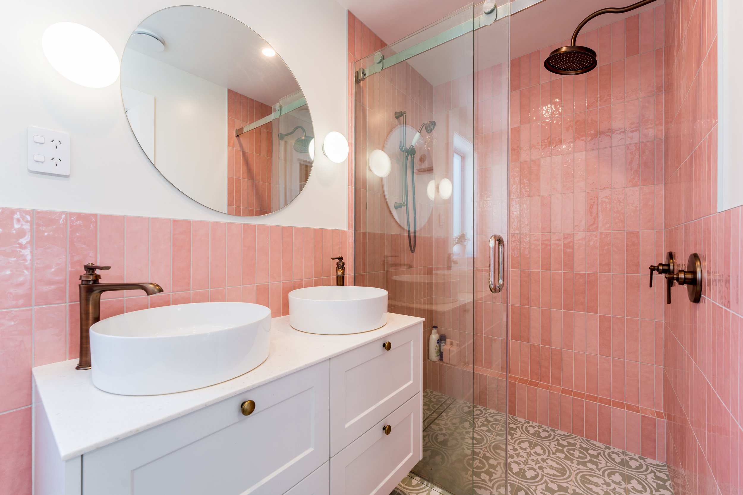 Bathroom with pink tiled shower and wall, white vanity with two oval vessel sinks and bronze fixtures, round mirror, glass shower door, patterned floor tiles, and small window.