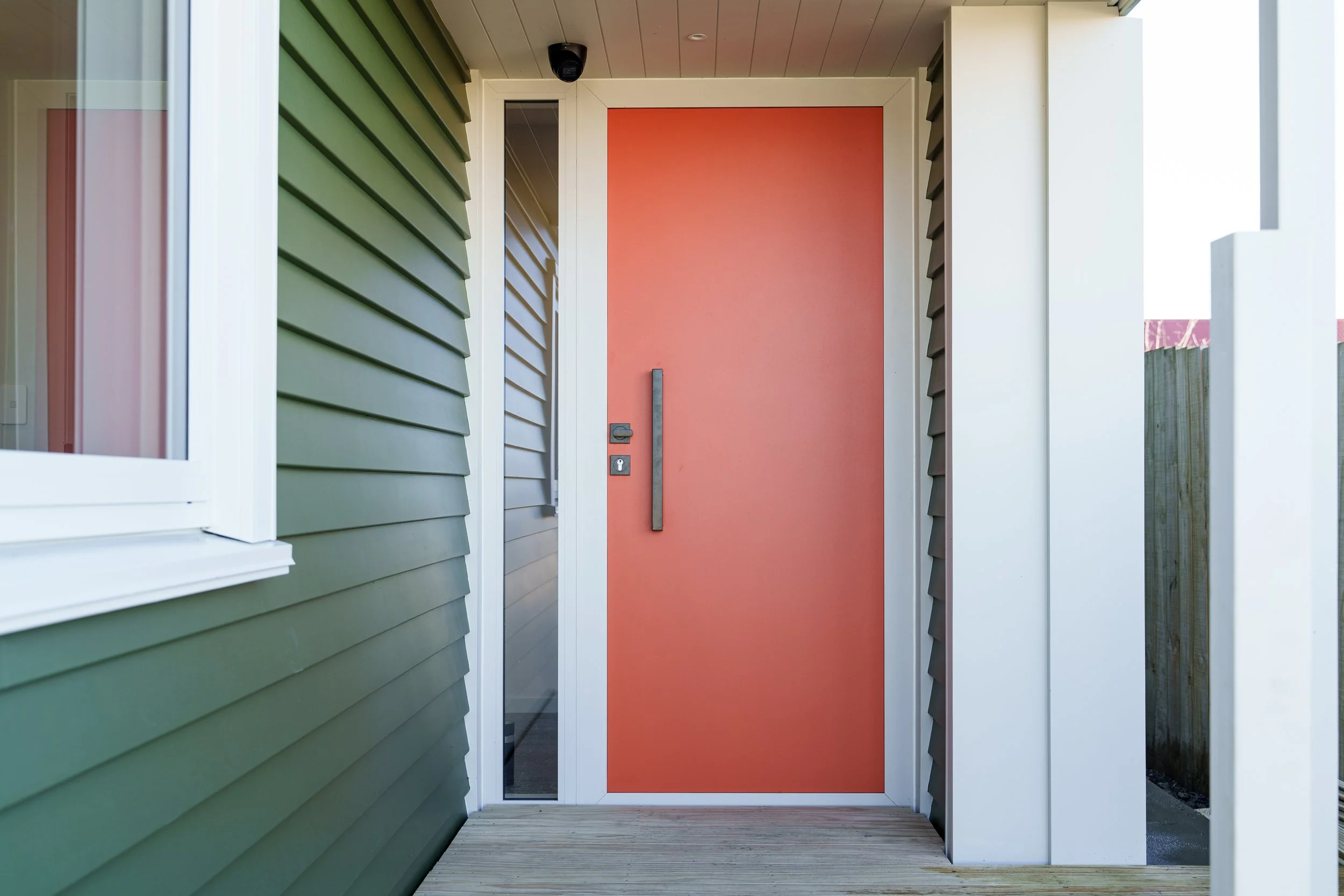 Close-up of an outdoor entry door painted in coral color with a vertical black handle and a keyhole. The door is framed by white trim, and a glass side panel is partially visible on the left. The exterior siding of the house features green horizontal