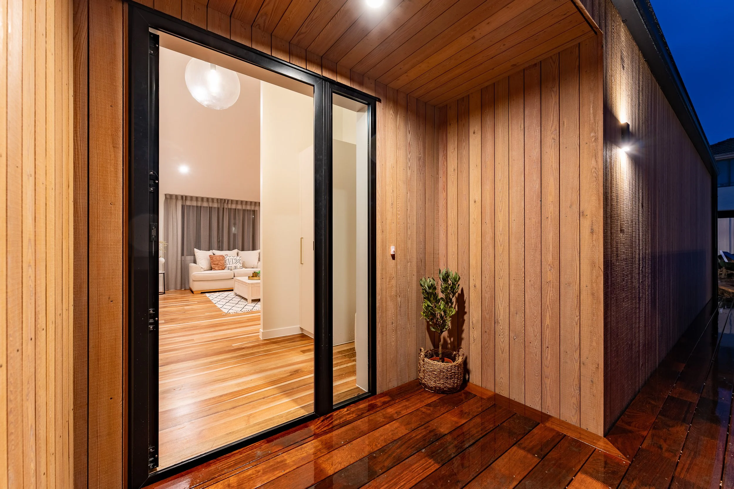 Passive house principles, high performing, warm and dry. View of a modern house exterior at night, showing a sliding glass door opening into a brightly lit living room with wooden flooring and furniture. Timber cladding