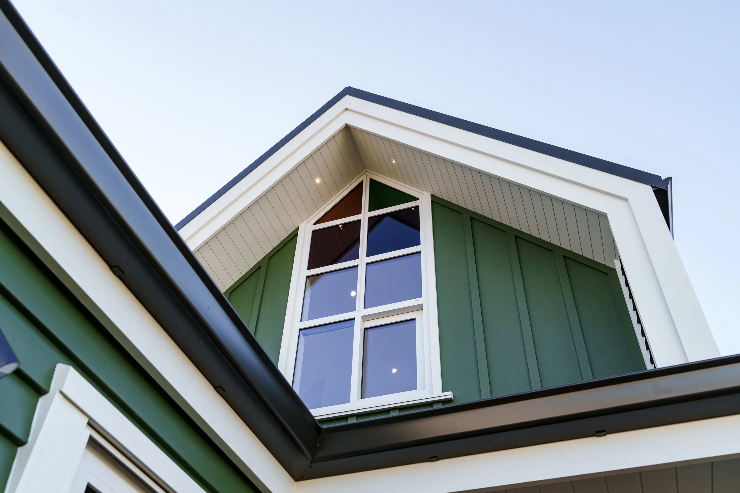 Passive house principles, high performing, warm and dry. Close-up look at the corner of a modern house with green siding, white trim, and a large multi-pane window, with a clear sky in the background. Solar power.
