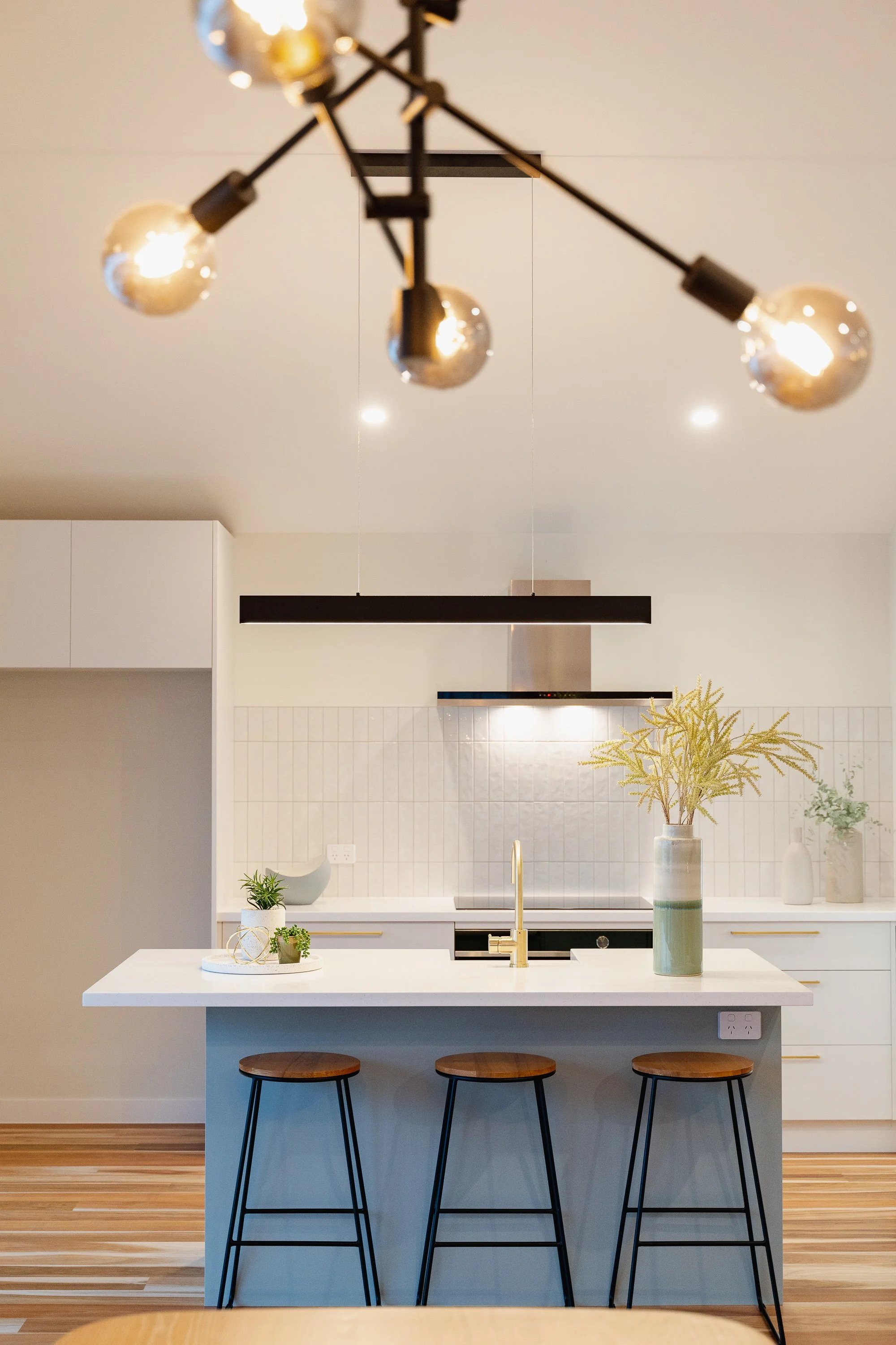 Modern kitchen with a white island, three wooden bar stools with black metal legs, a gold faucet, a white backsplash, and decorative plants in vases. A black ceiling light fixture is visible.