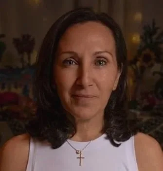 A woman with shoulder-length dark hair wearing a white sleeveless top and a gold cross necklace, standing indoors with a blurred floral background.