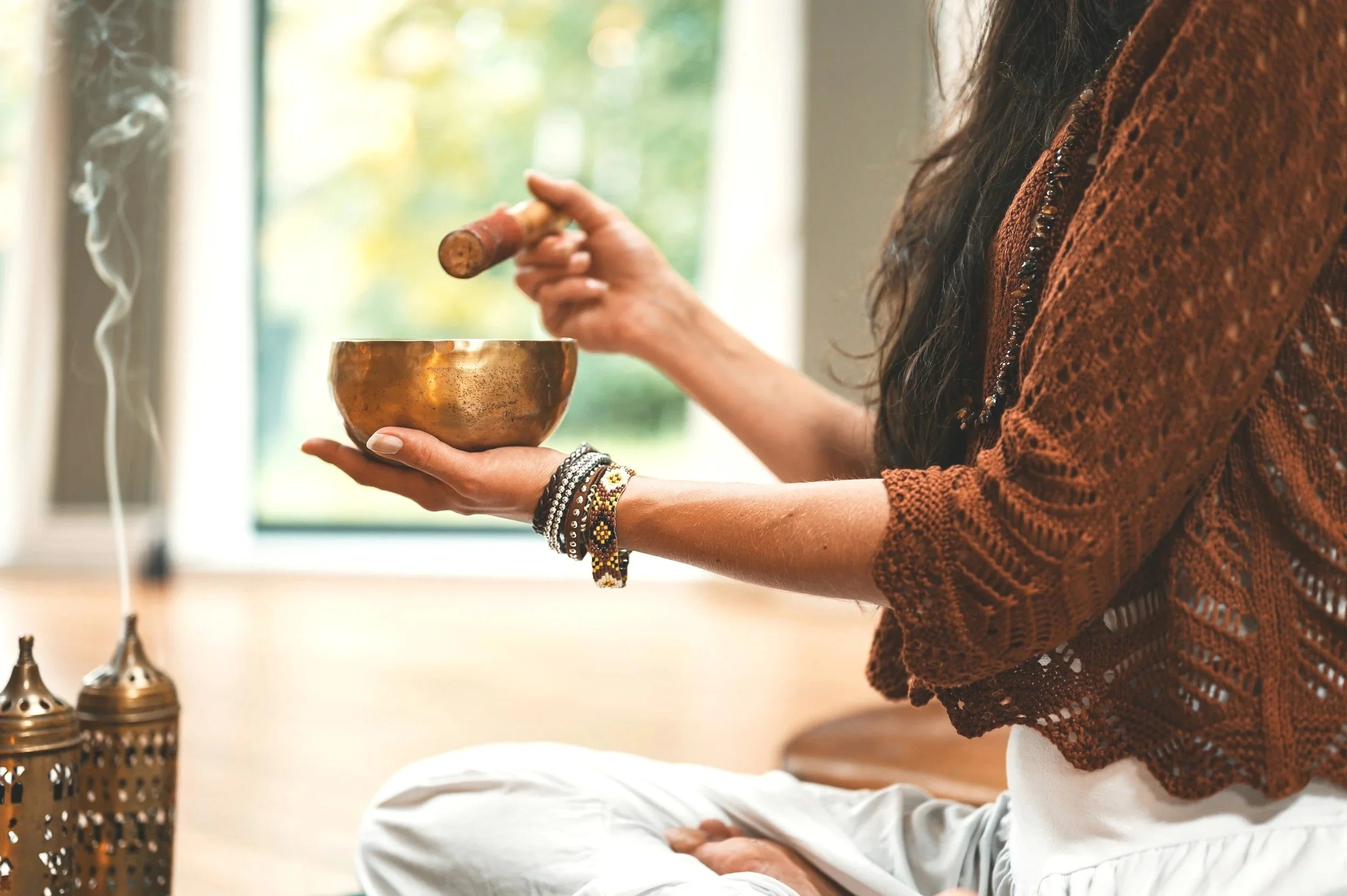 A woman sitting cross-legged indoors, holding a singing bowl in her left hand and a wooden mallet in her right hand, with steam rising from incense or candles on the side and a window showing greenery outside.