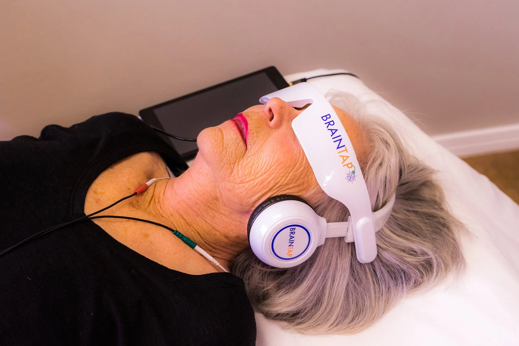 An elderly woman with gray hair lying on a bed, wearing headphones labeled 'BRAIN TAP' and a visor with the same logo, participating in a brain stimulation or therapy session.