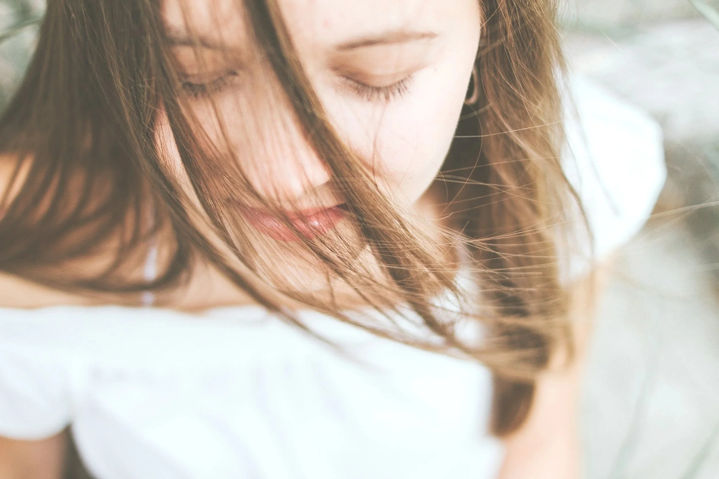 Close-up of a woman with long brown hair and closed eyes, wearing a white shirt.