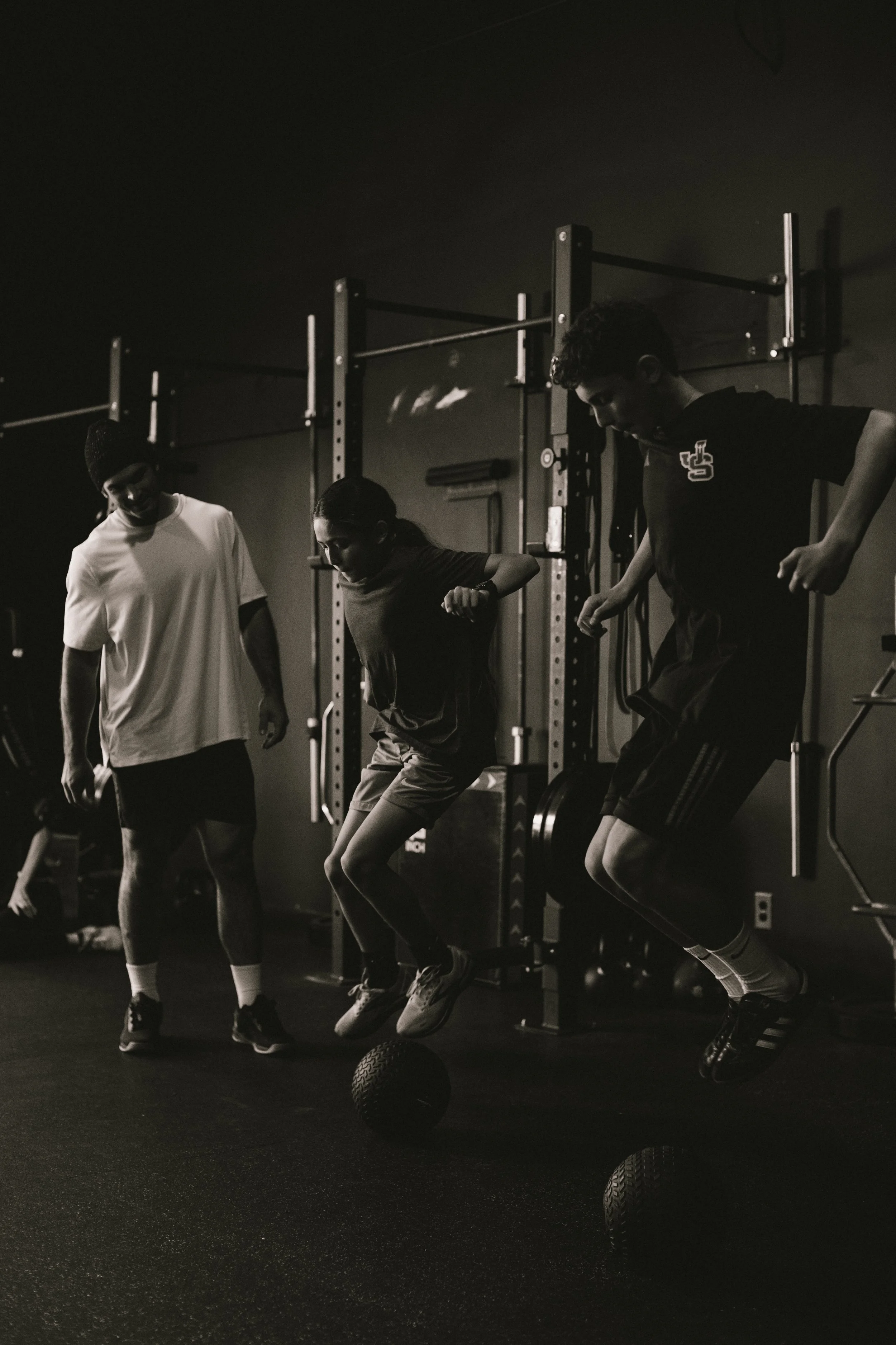 Three young people jumping in a gym with black medicine balls on the floor; two are mid-air, one is standing, all appear to be participating in a fitness activity.