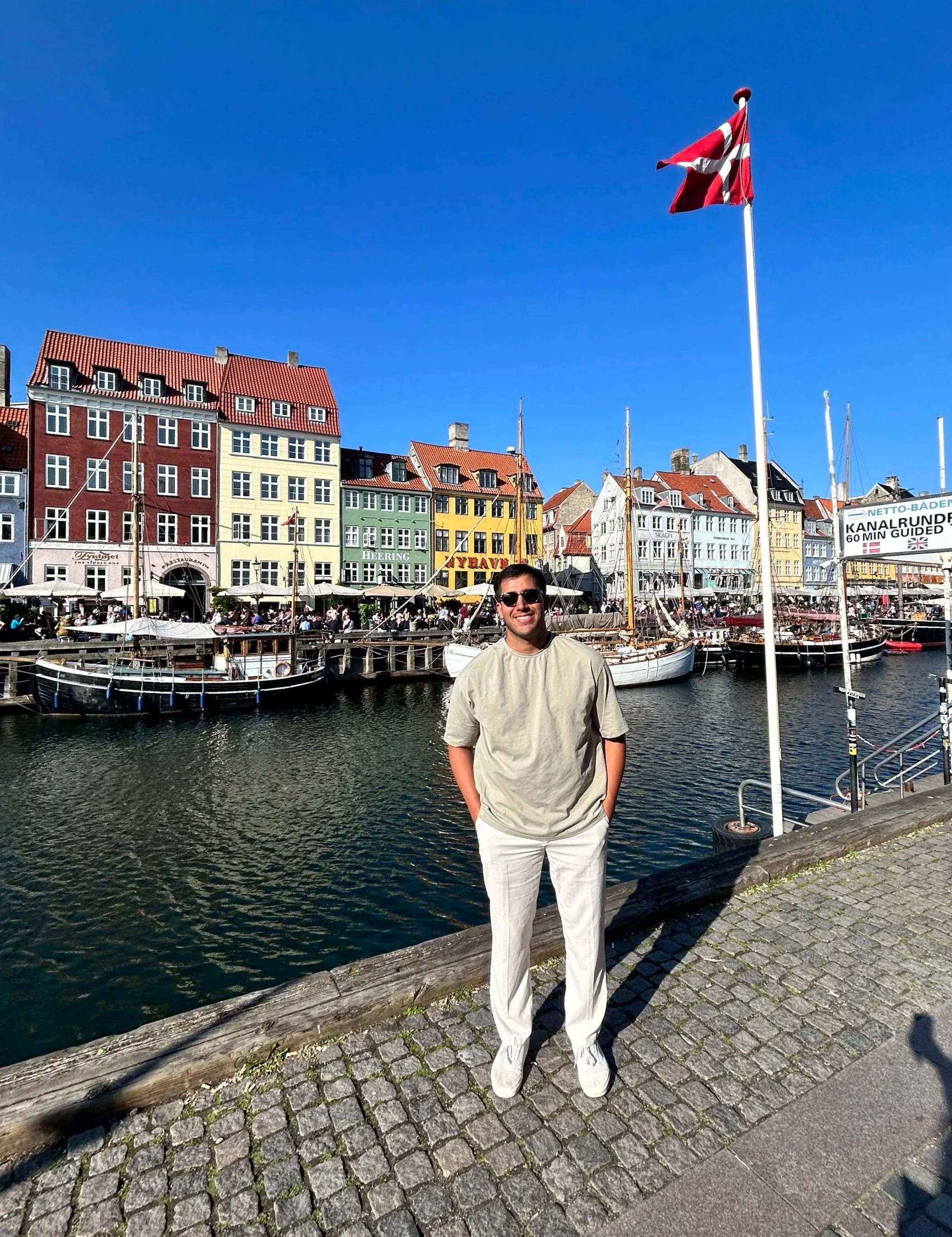 A man wearing sunglasses, a beige shirt, and white pants standing on cobblestone beside a canal with colorful buildings and boats in the background.