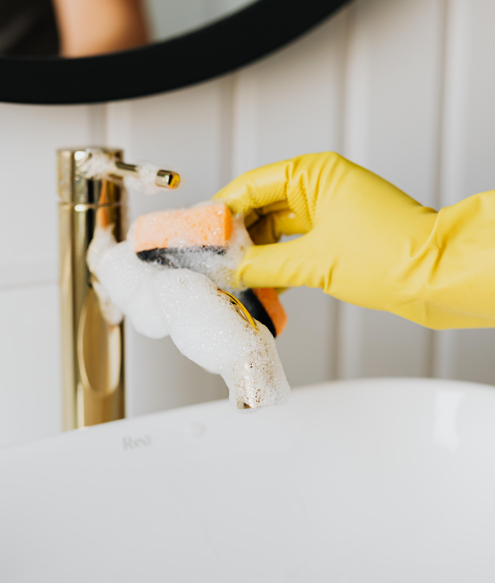 Person wearing yellow rubber gloves washing a sink faucet with soap and a sponge.