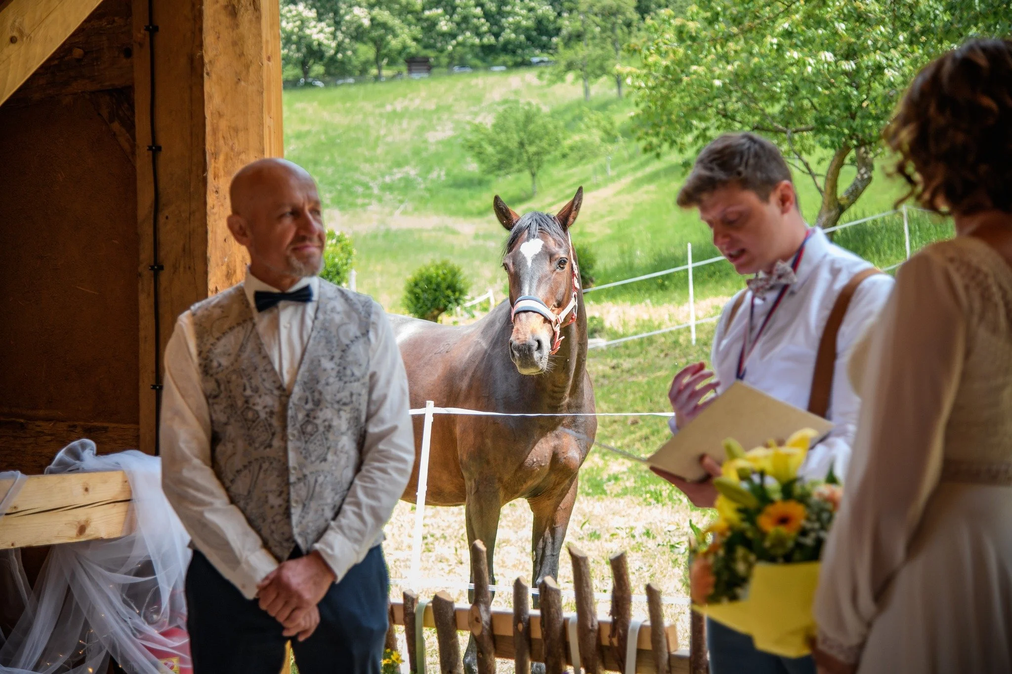 Lidé oblečeni ve formálním a vintage oblečení v koňské stáji během svatby, s koněm v pozadí.