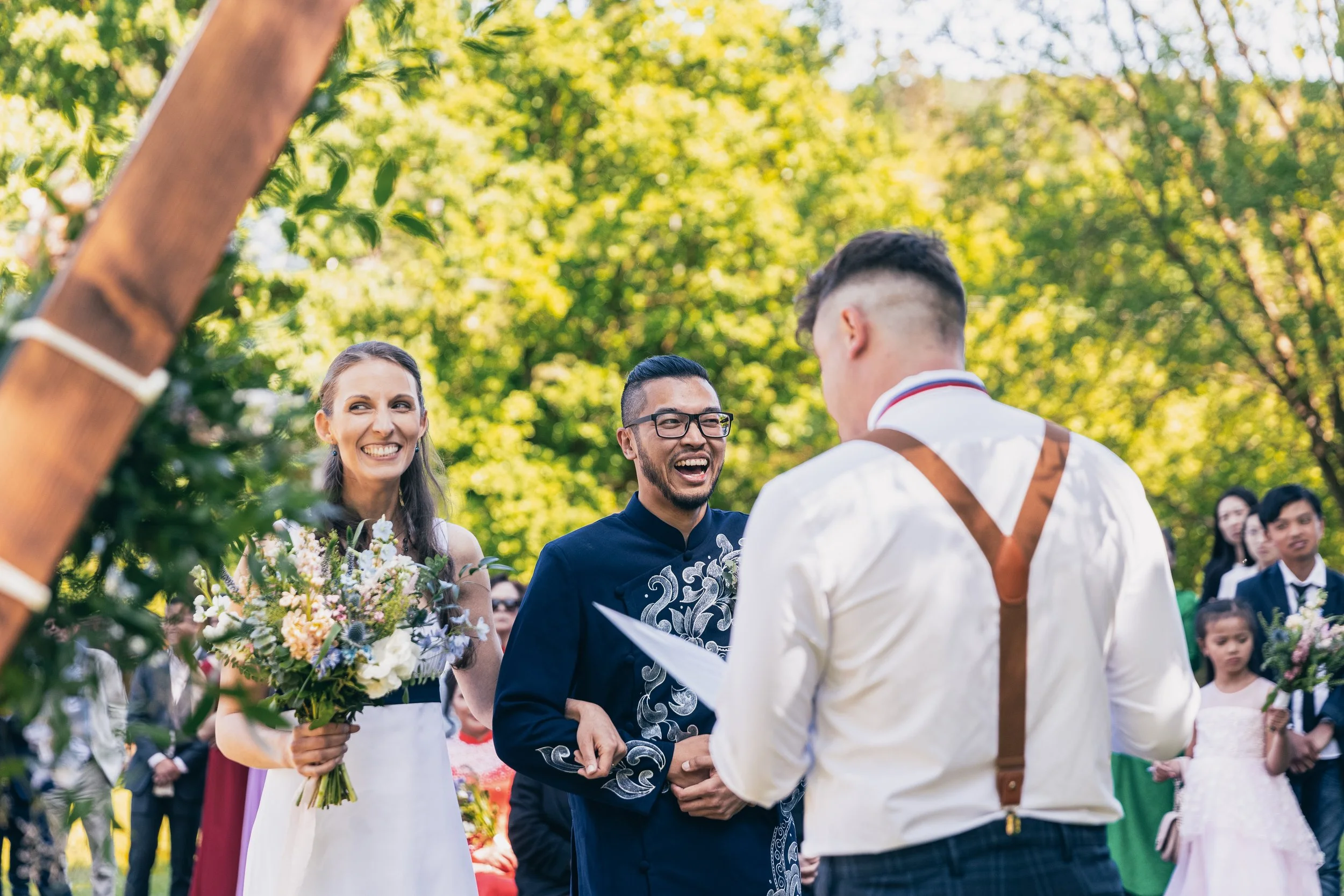 Svatební obřad venku s nevěstou, ženichem a oddávajícím, obklopeni hosty a svěžími zelenými stromy.