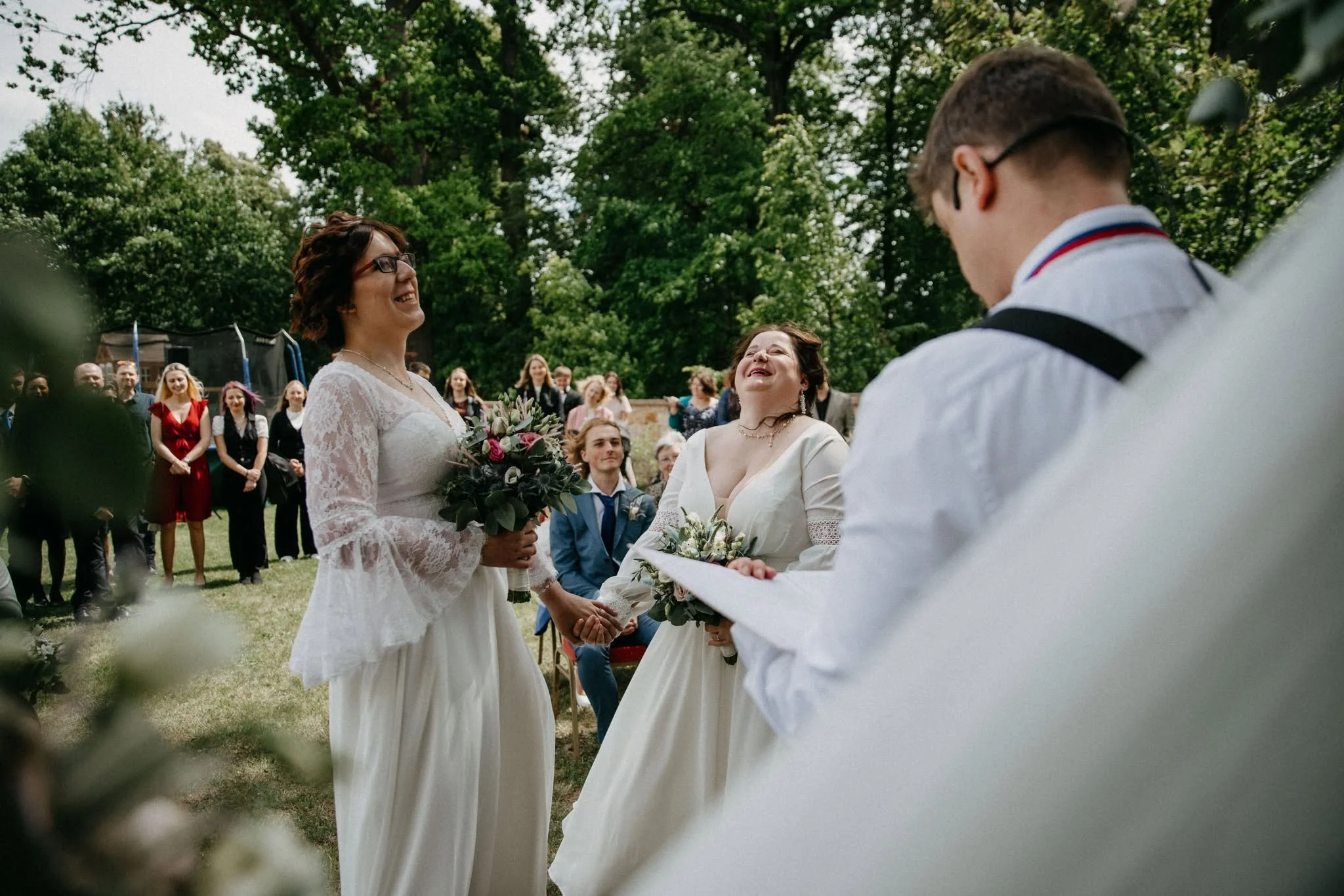 Dvě ženy ve svatebních šatech se drží za ruce během venkovního svatebního obřadu, usmívají se a smějí se, s mužem, který vede svatební obřad, obklopené hosty a zelení.