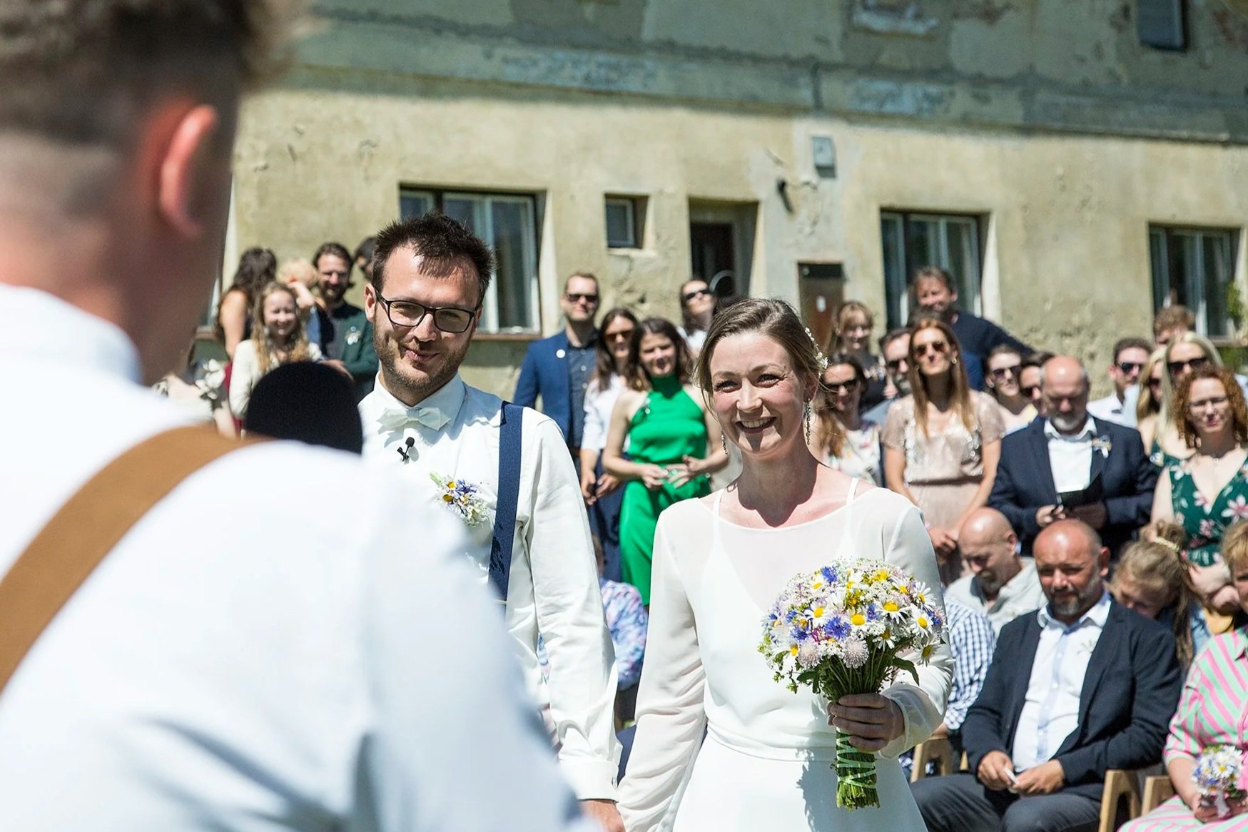 Nevěsta drží kytici květin a usmívá se na svého ženicha během svatebního obřadu venku, v pozadí sedí a stojí hosté.