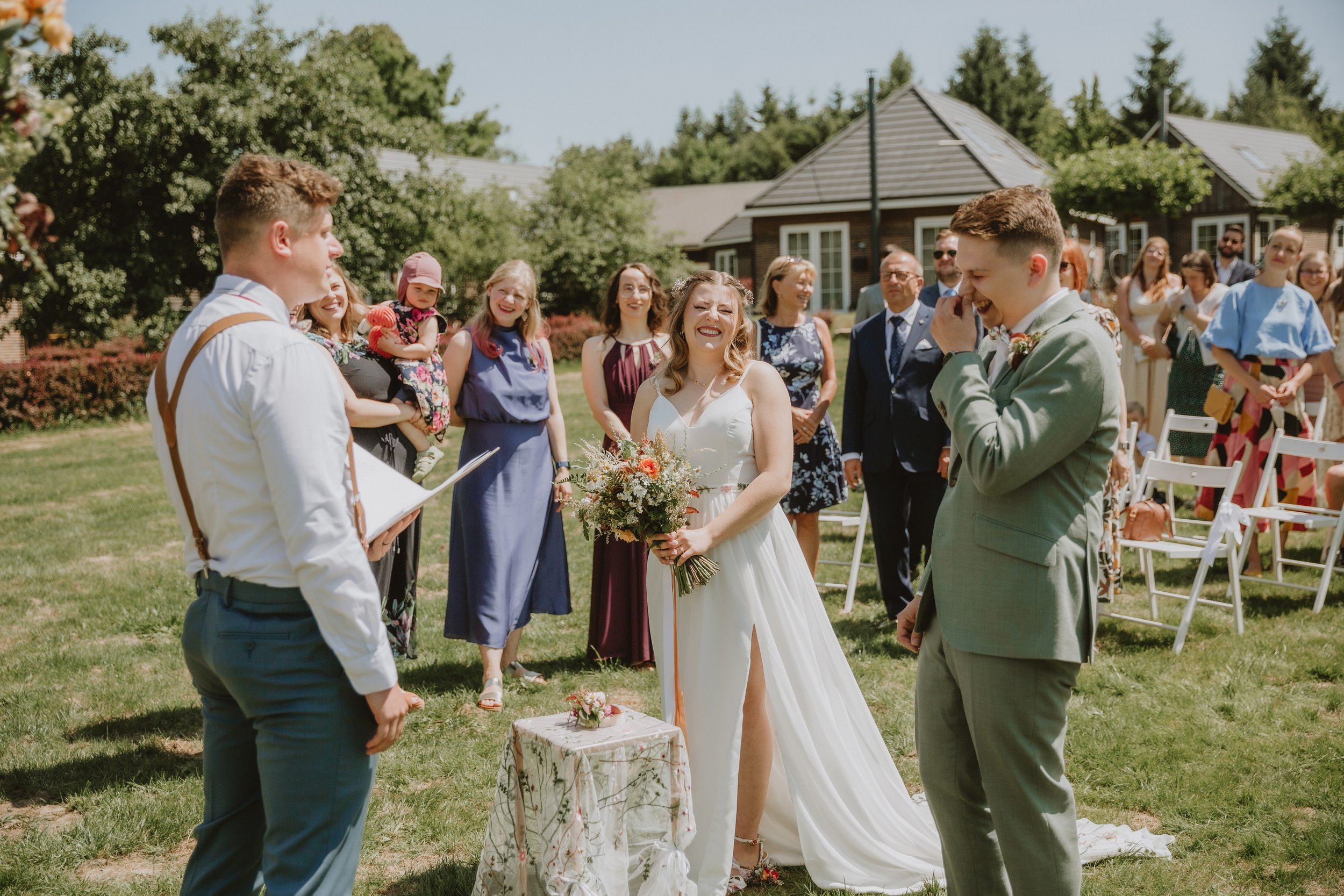 Nevěsta v bílých svatebních šatech držící kytici v rukou se usmívá na svého ženicha během venkovního svatebního obřadu. Ženich má na sobě světle šedý oblek, usmívá se a zakrývá si ústa. V pozadí stojí hosté a pozorují pár.