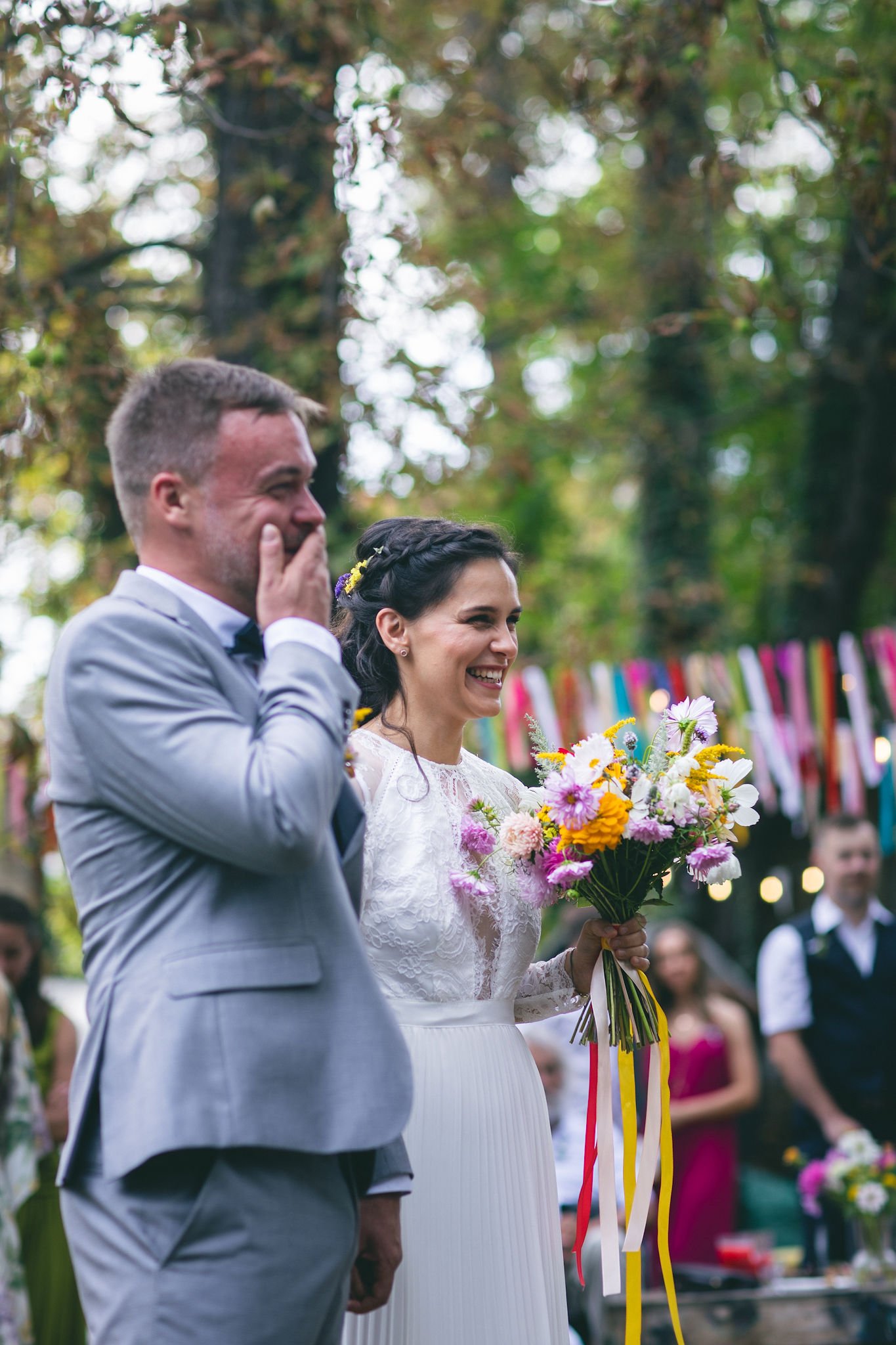 Usmívající se nevěsta v bílých šatech držící barevnou kytici květin, stojící vedle ženicha v šedém obleku, na venkovním svatebním obřadu obklopená stromy a hosty.