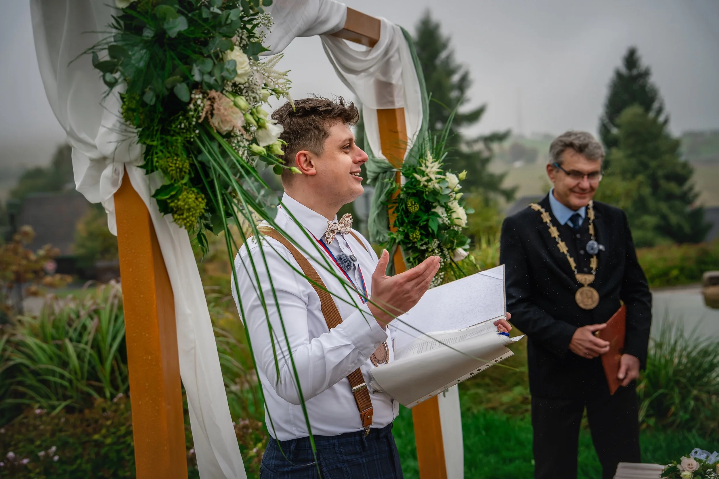 Mladý muž v bílé košili a motýlku čte svatební sliby venku během svatebního obřadu, poblíž stojí oddávající v černém obleku a brýlích, oba pod zdobeným obloukem s květinami a zelení, za deště.
