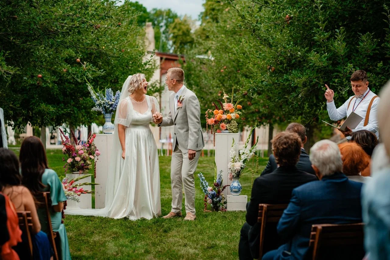 Svatební obřad venku, nevěsta a ženich se drží za ruce, usmívají se, obklopeni barevnými květinovými aranžmá a hosté se dívají. Obřad se koná pod stromy na trávníku.