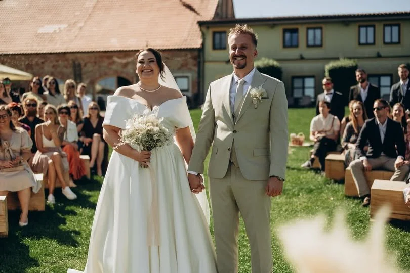 Nevěsta a ženich se drží za ruce během venkovního svatebního obřadu, obklopeni sedícími hosty na trávě, před domem a stodolou, slunečný den.
